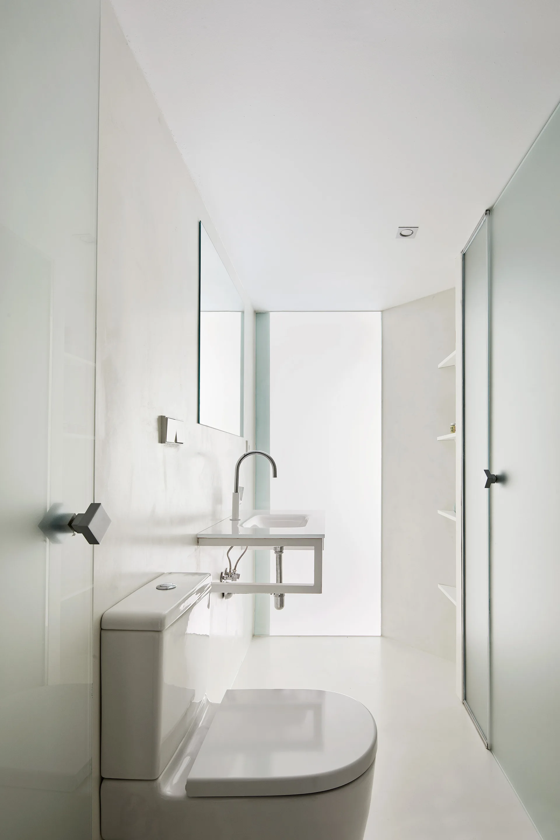 A narrow minimalist white bathroom with a wall-mounted basin, rectangular mirror, toilet, and a frosted glass shower door.