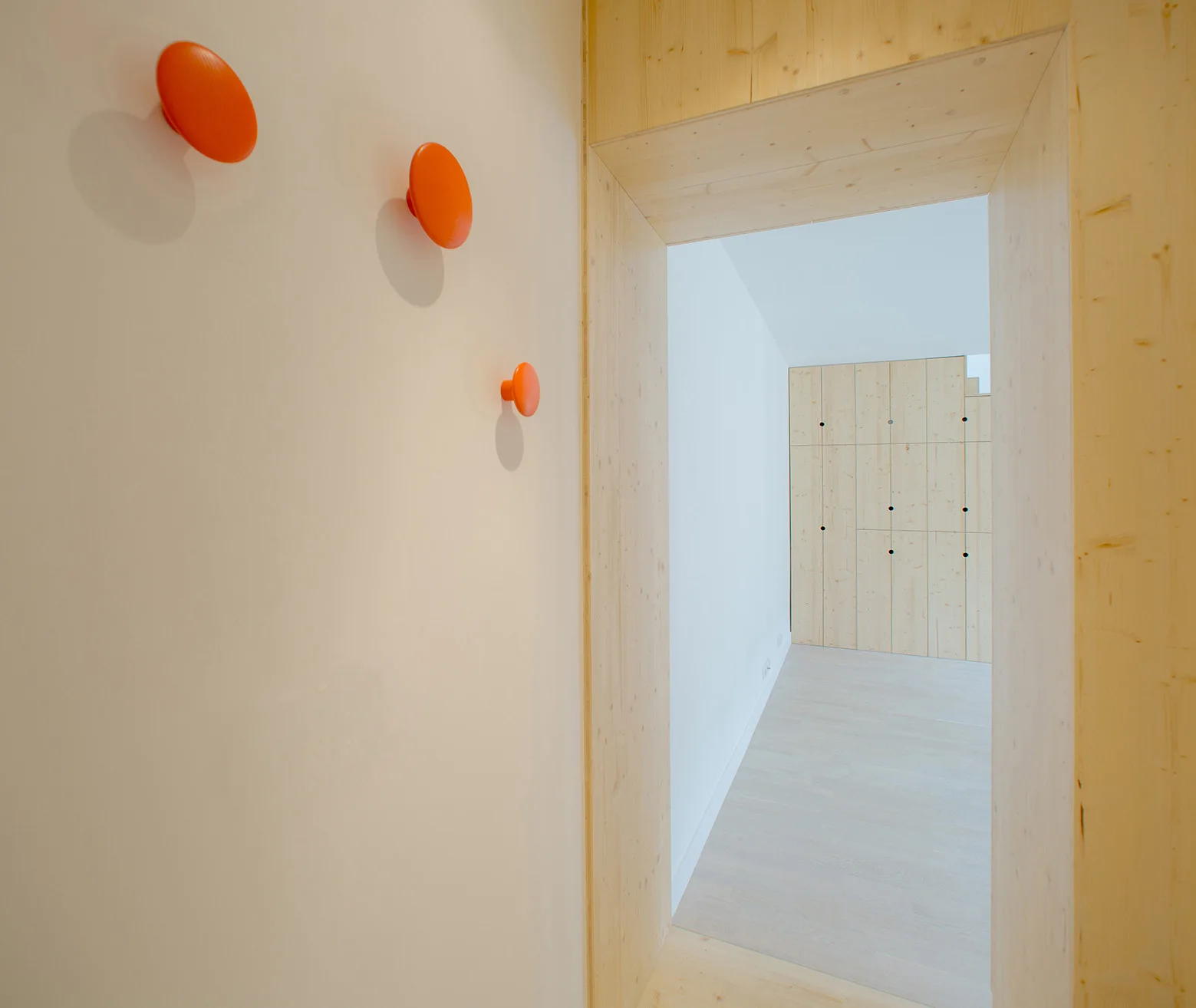Three round orange hooks on a white wall lead to a wide light wood doorway framing a built-in wooden storage cabinet in a minimalist hallway.