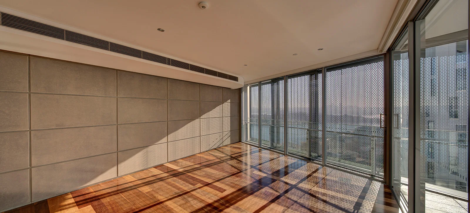 An empty minimalist room featuring textured grey wall panels, polished hardwood floors, and sliding glass doors covered by perforated metal privacy screens.