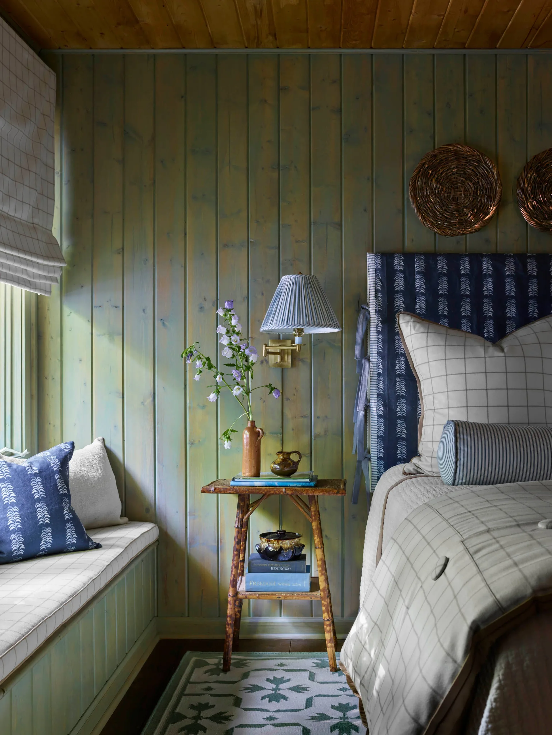A cozy bedroom corner features green stained wood walls, a small bamboo side table, a built-in window bench, and a bed with checkered and striped linens.