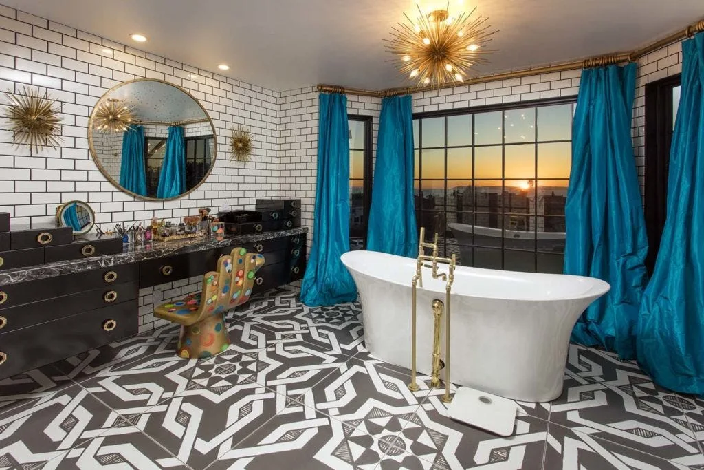 A bathroom featuring a freestanding white bathtub, bold geometric floor tiles, white subway tile walls, and vibrant blue curtains framing a window.