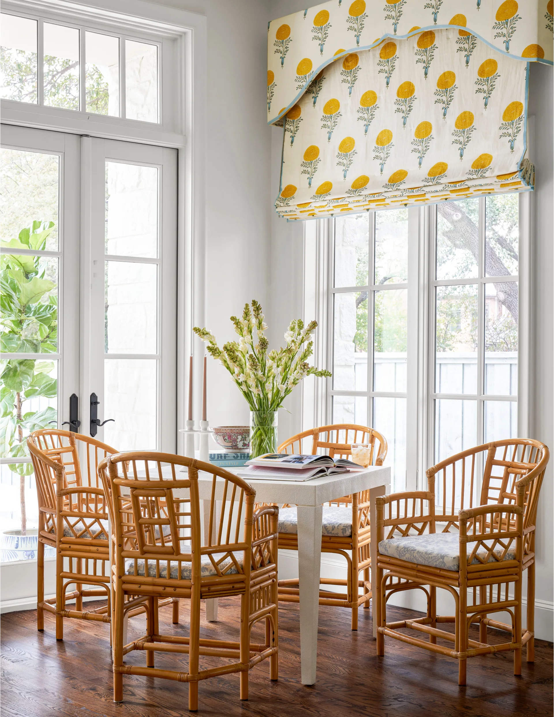 A white square dining table surrounded by four bamboo chairs sits near large windows with a yellow floral valance and french doors.