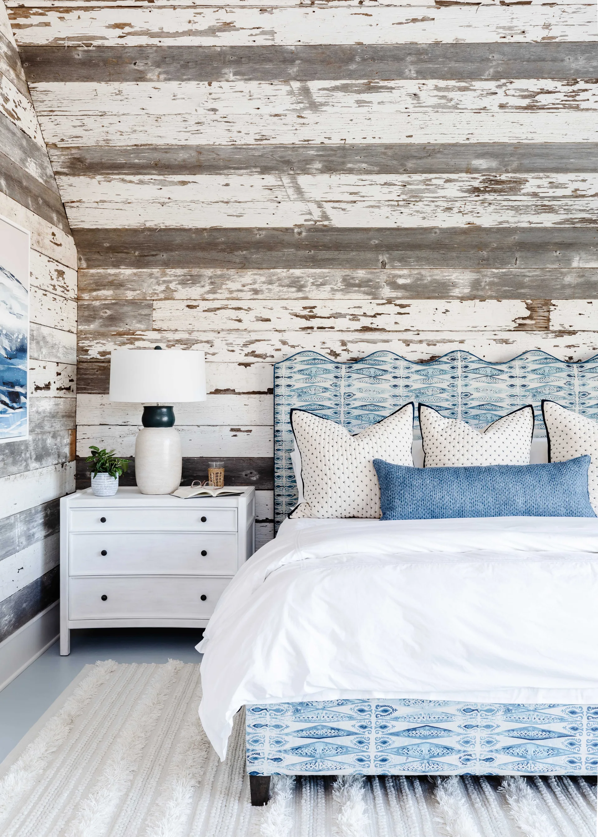 A bedroom features an upholstered blue patterned bed against a rustic distressed wood plank wall alongside a white three-drawer nightstand.