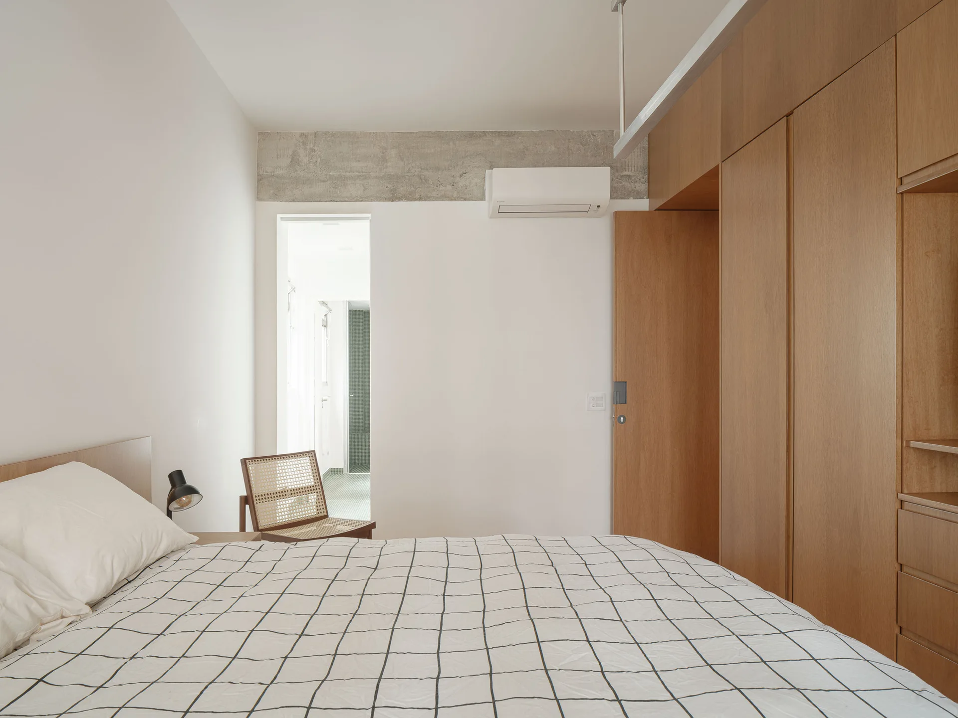 A minimalist bedroom featuring warm wood wardrobes, a bed with black and white grid bedding, and an exposed concrete beam.