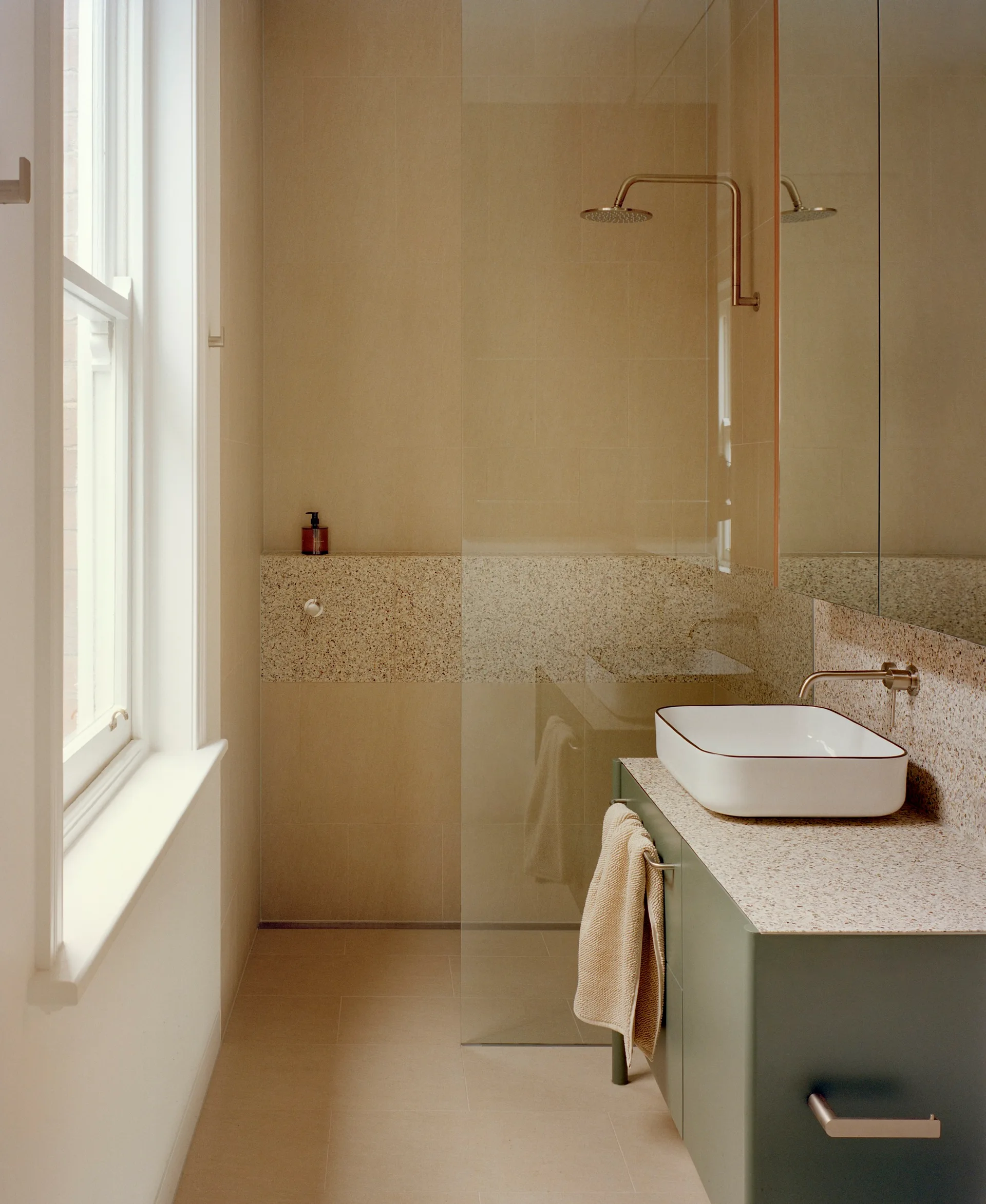 A modern bathroom with a muted green vanity, white vessel sink, a walk-in shower with a glass panel, beige tiles, and speckled stone counters.