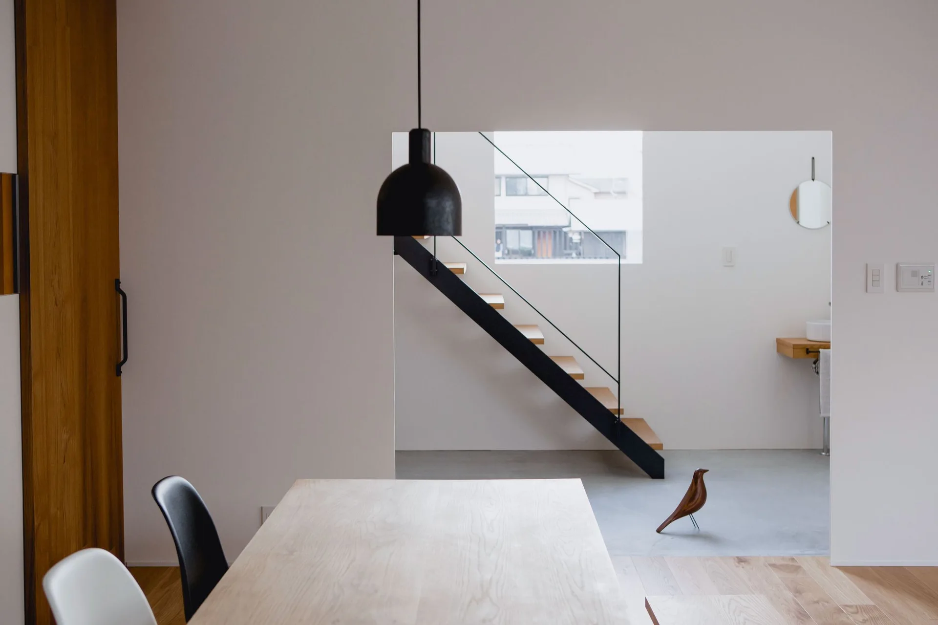 A minimalist dining area with a light wood table and black pendant light leads to an open-riser staircase with wood treads and a black metal stringer.
