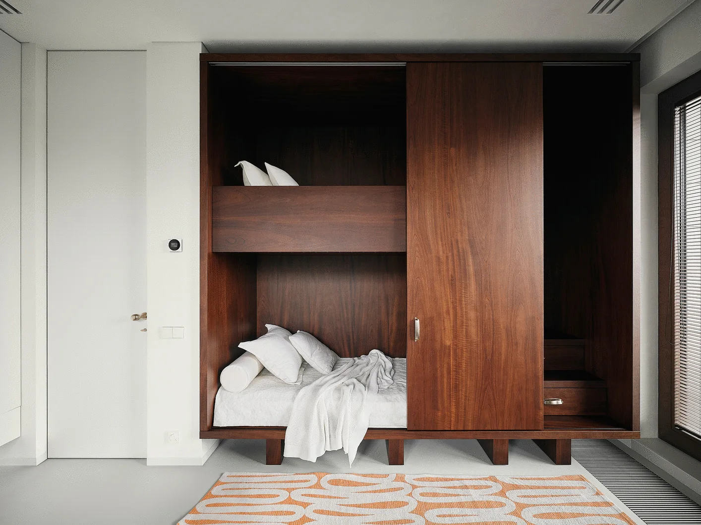 A dark wood built-in wardrobe features an integrated bed nook with white bedding next to an orange and white patterned rug.