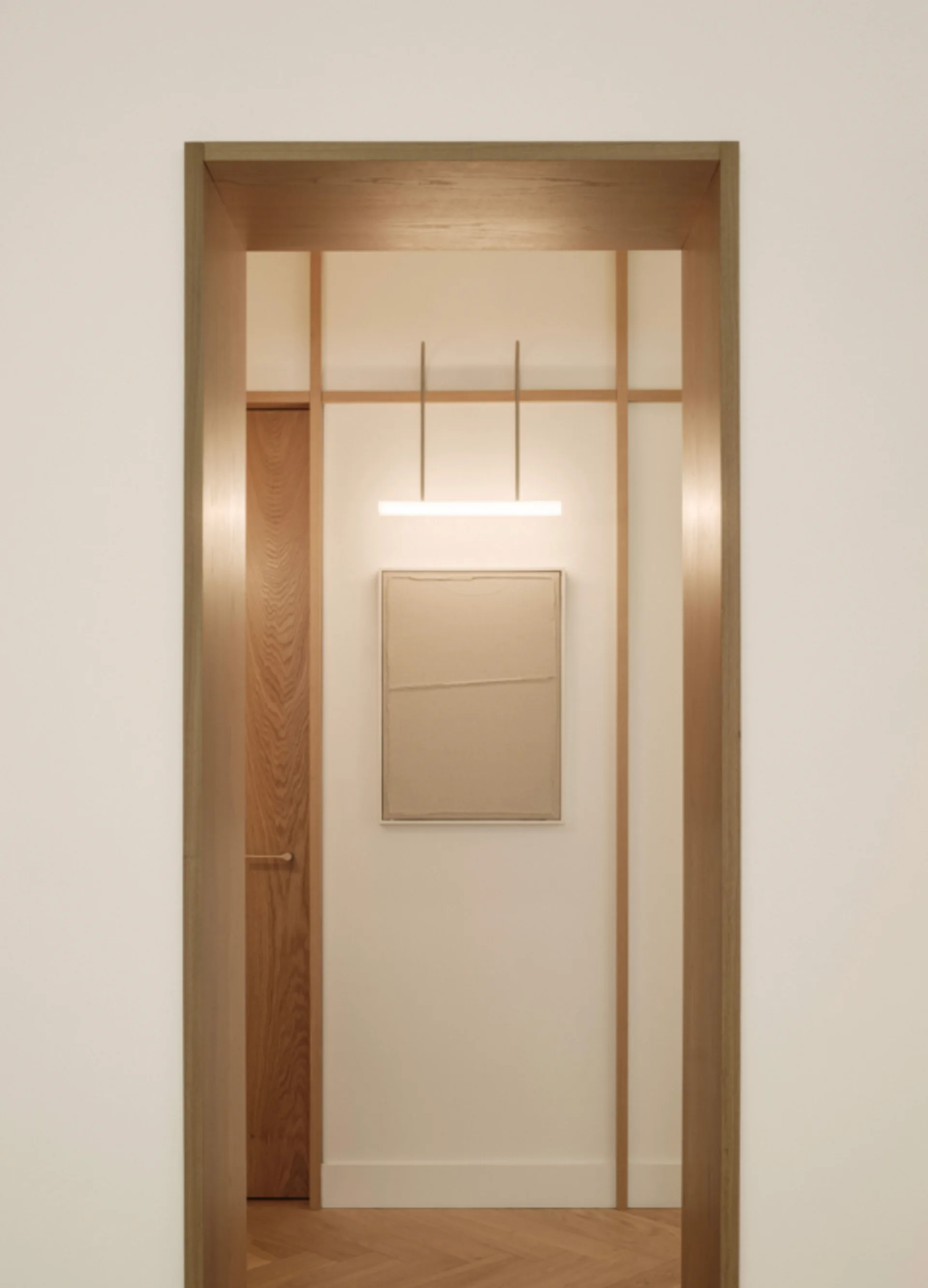A view through a wood-framed doorway into a minimalist corridor with a linear light, vertical wood wall trim, and textured framed artwork.