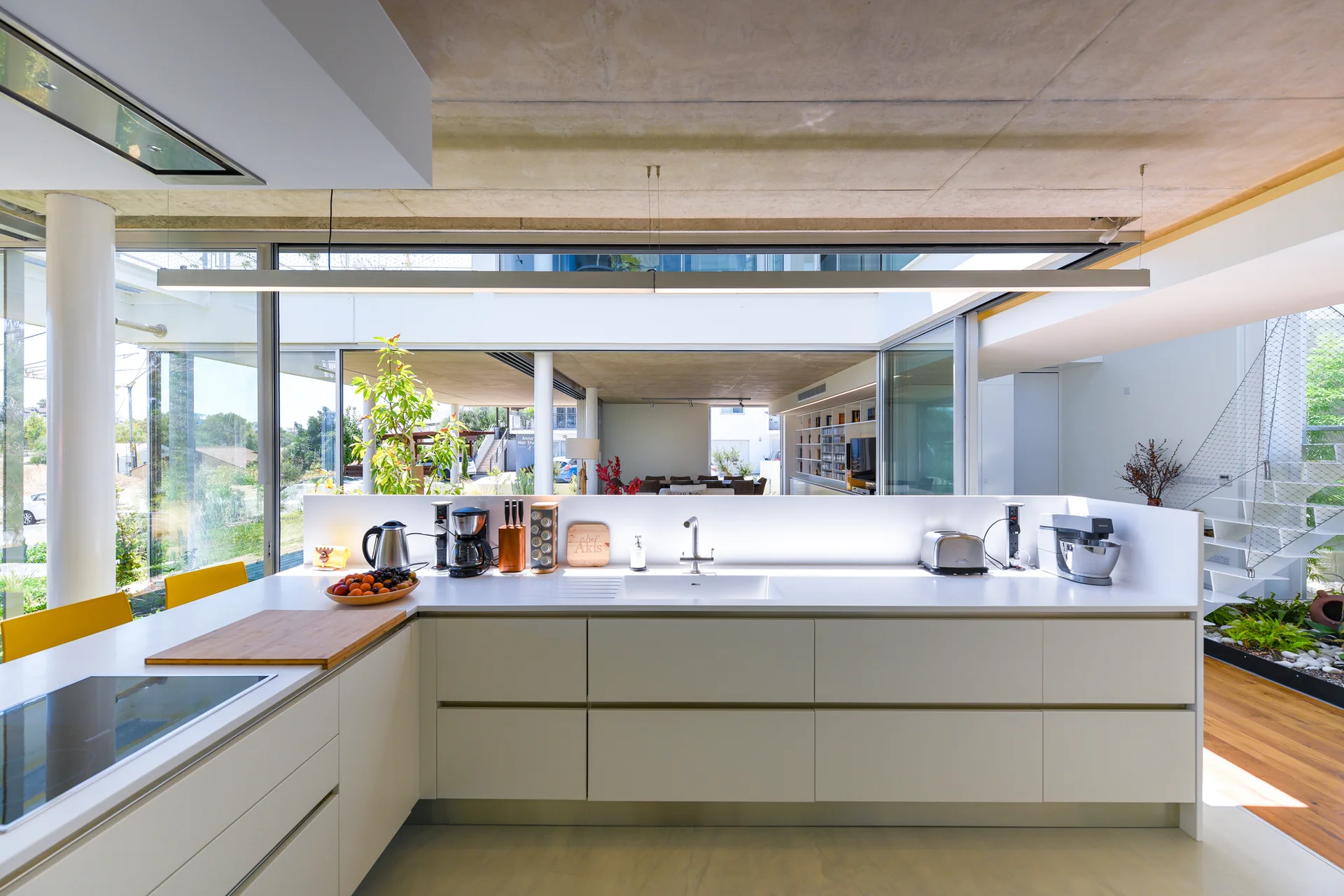 Minimalist white kitchen features flat-panel cabinets, a glowing low backsplash, a concrete ceiling, and views of a courtyard through glass walls.