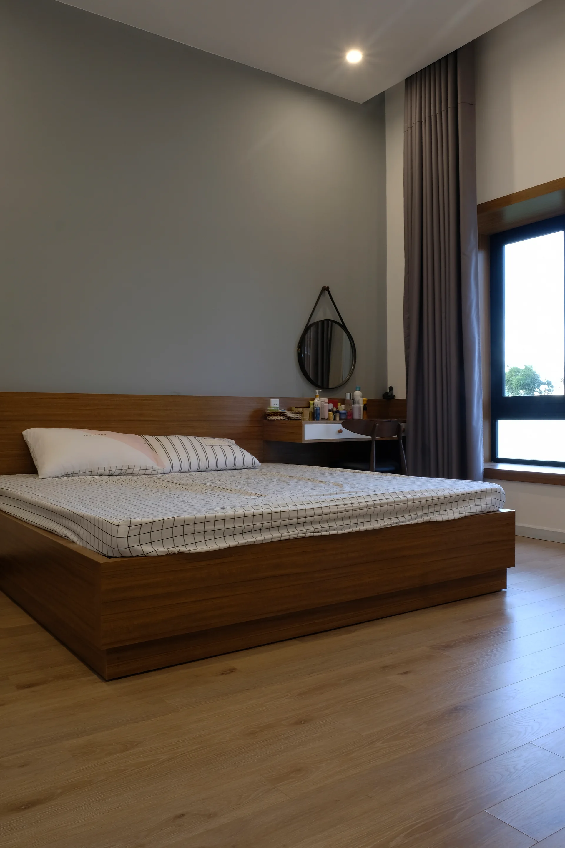 A contemporary bedroom featuring a low wooden platform bed, grey walls, and a floating wooden vanity desk with a round hanging mirror.