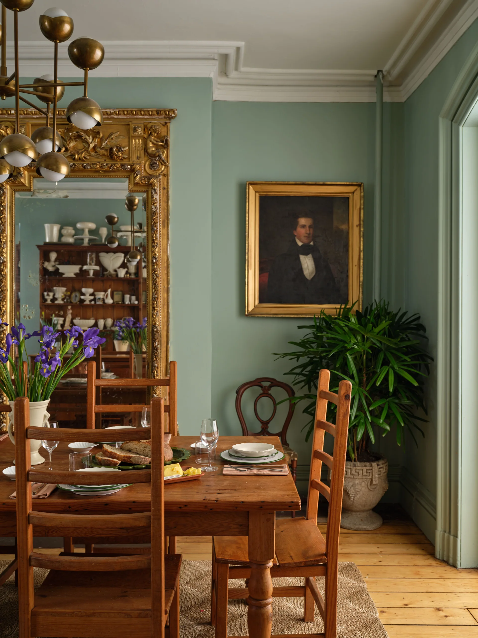 A traditional dining room featuring sage green walls, a rustic wooden table with ladder-back chairs, and a large ornate gold-framed mirror.
