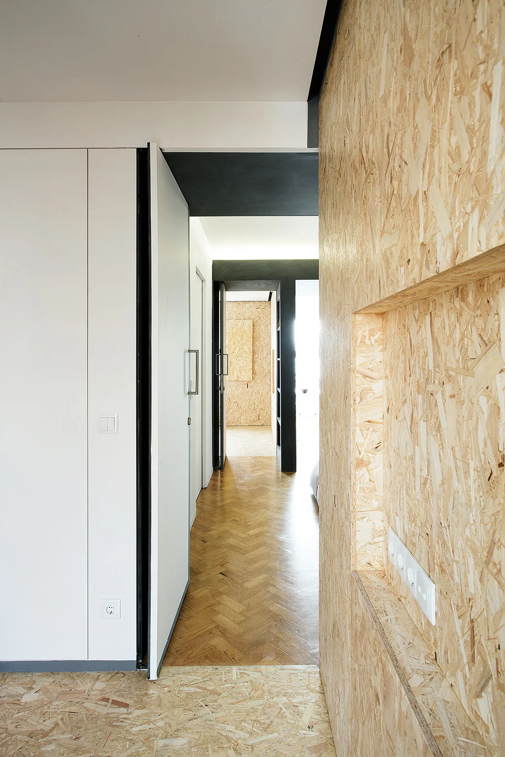 An interior hallway showing raw OSB wood wall paneling with a recessed niche, a minimalist white door, and herringbone wood parquet flooring.