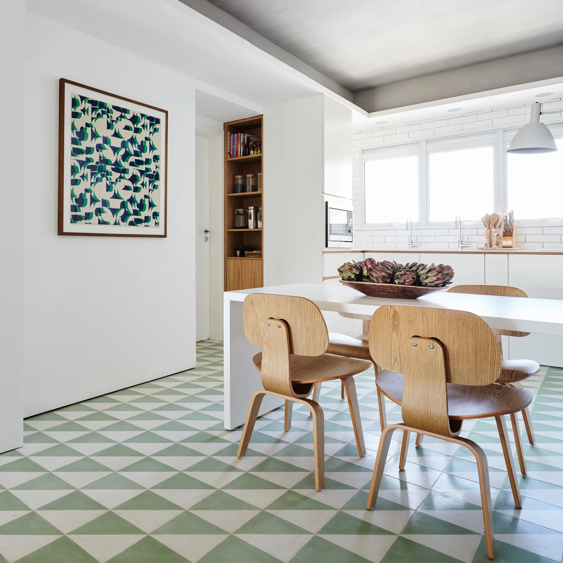 A modern dining table with molded plywood chairs sits on a green and white triangle-patterned tile floor next to a white wall with framed graphic art.