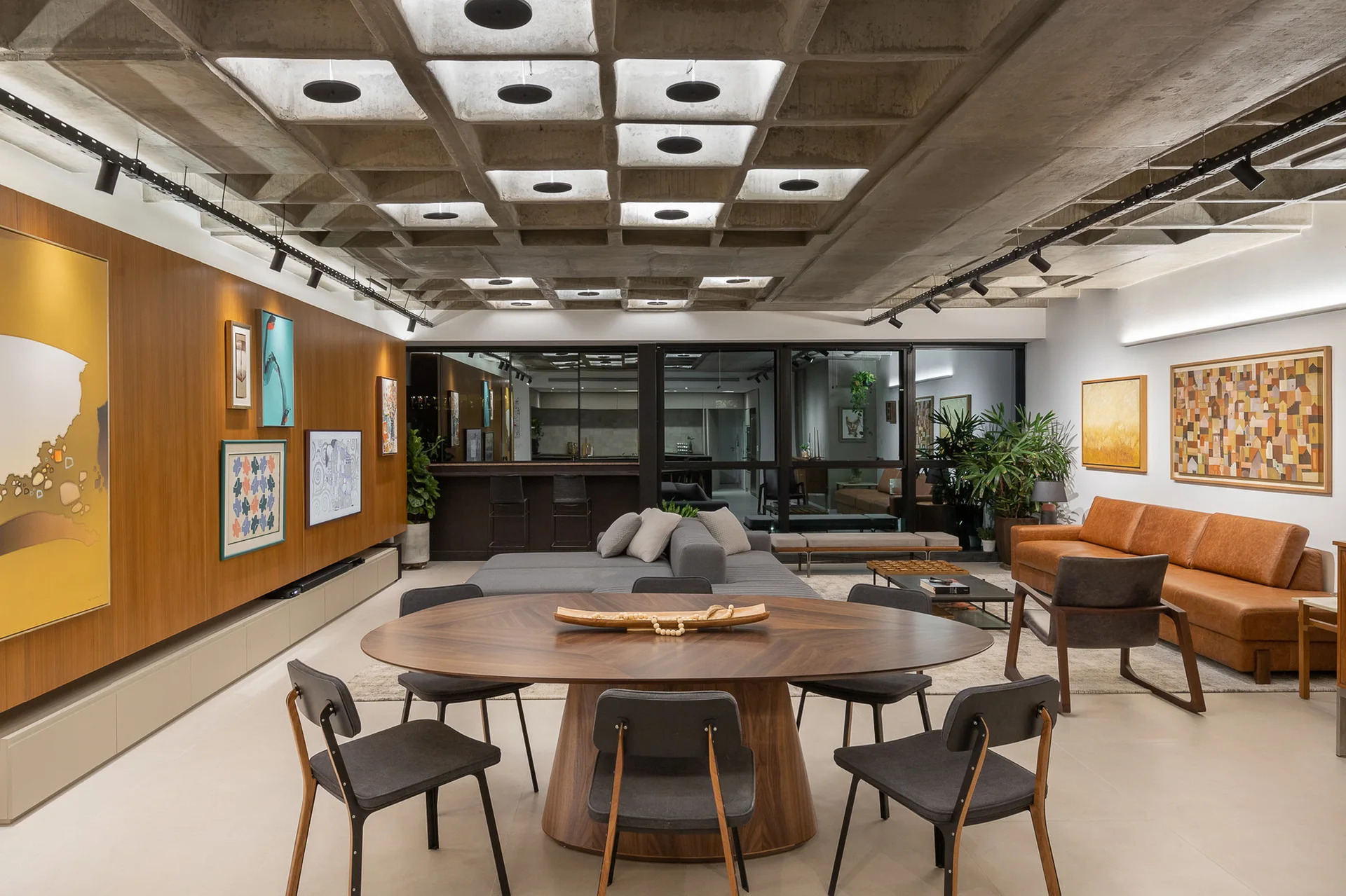 A modern open-concept dining and living space featuring a concrete waffle ceiling, a wood-paneled wall, and an oval wood dining table.