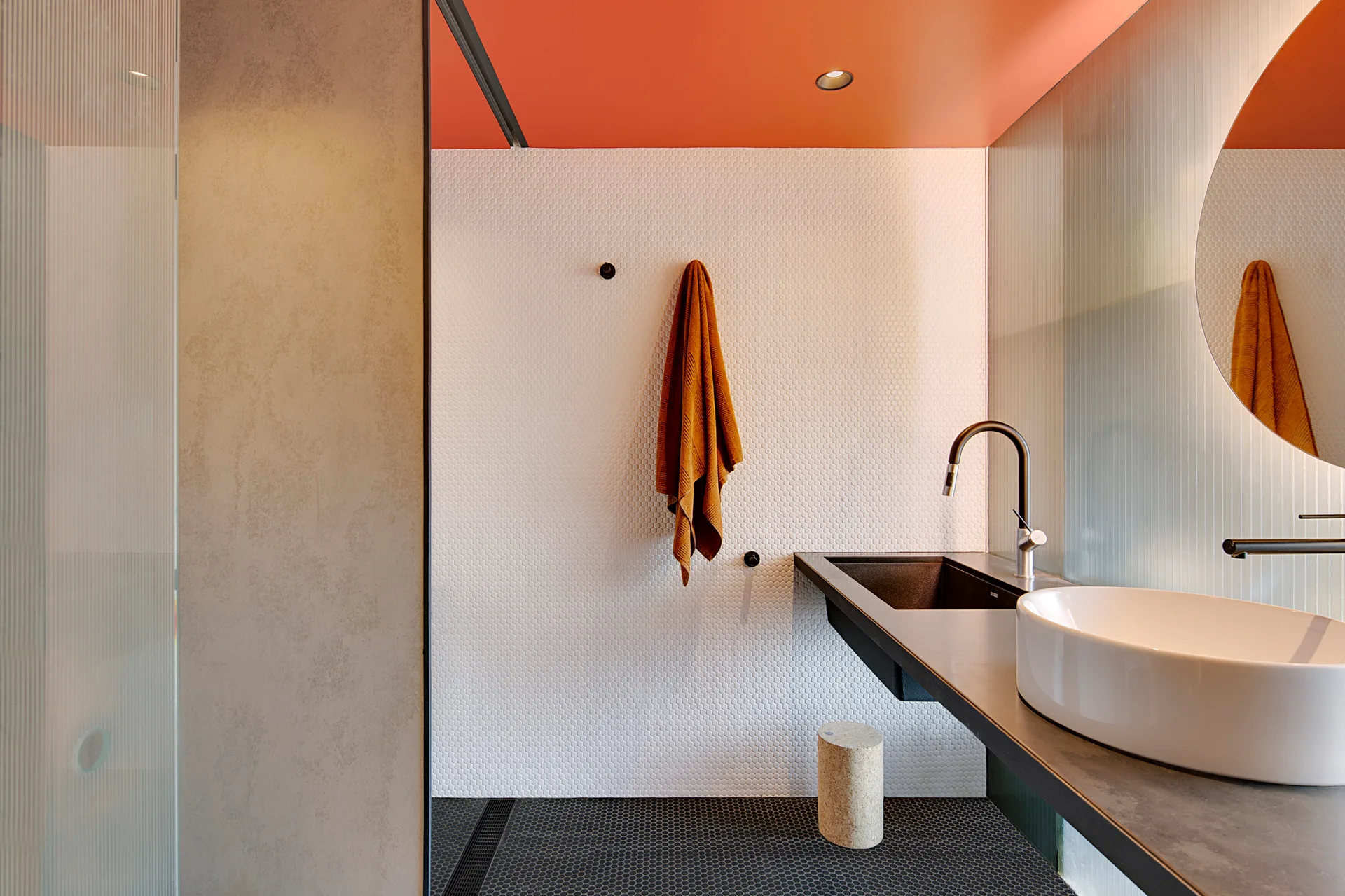 A modern bathroom with white penny tile walls, an orange ceiling, a dark floating vanity with a white vessel sink, and a circular mirror.
