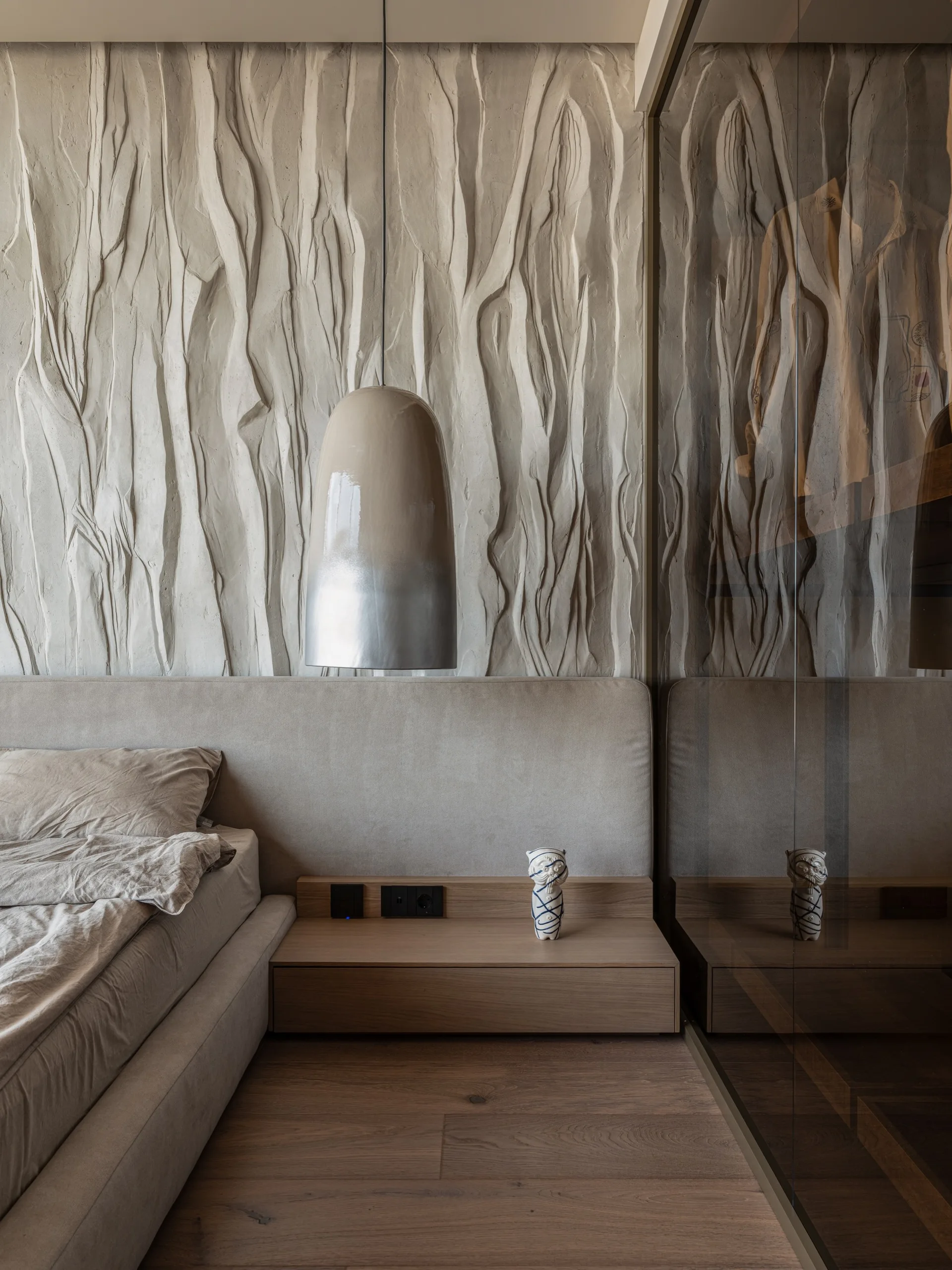 A low upholstered bed sits against a deeply textured plaster wall, flanked by a wooden nightstand and a metallic pendant light.