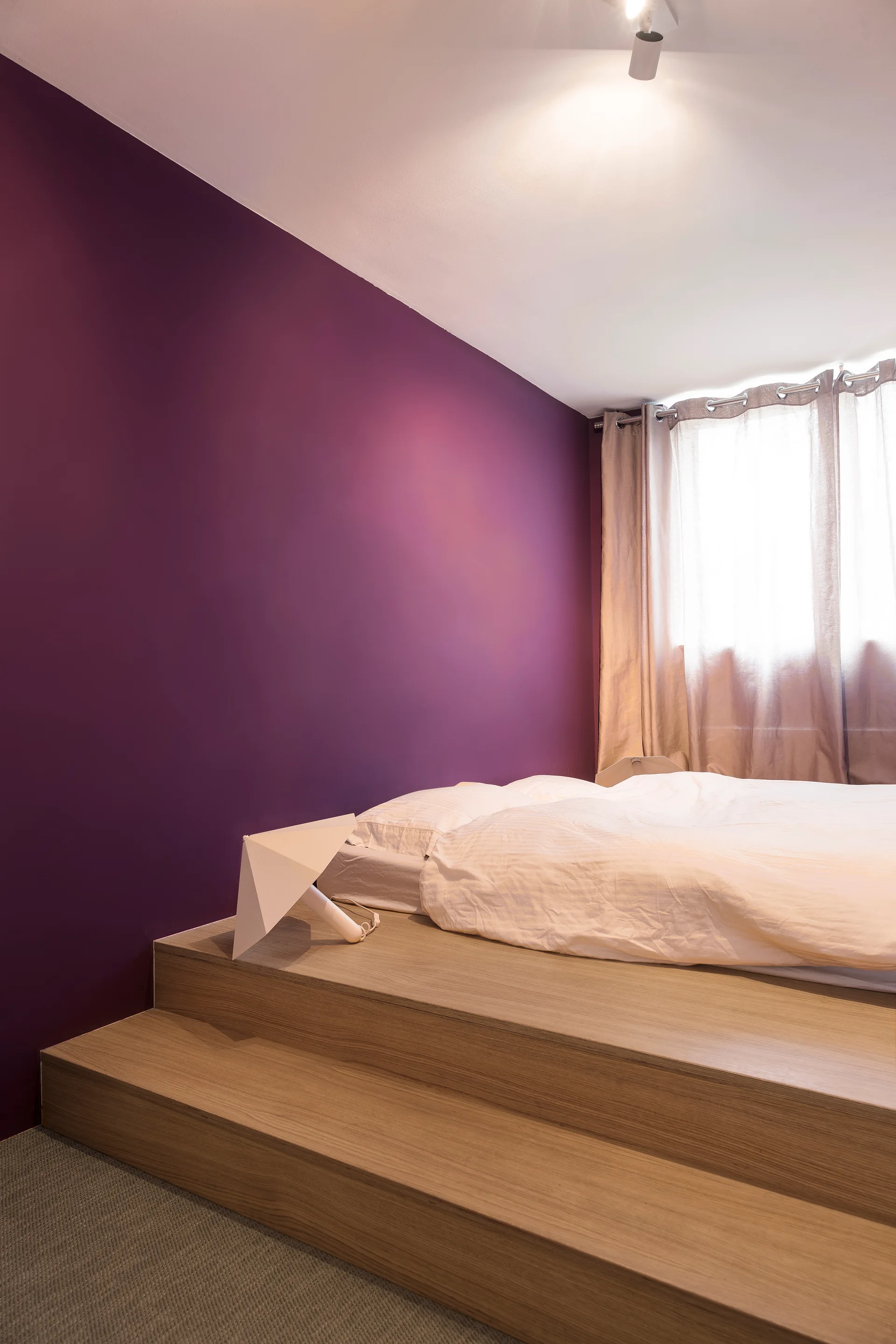 A minimalist bedroom featuring a bed on a stepped wood-toned platform set against a deep purple accent wall.