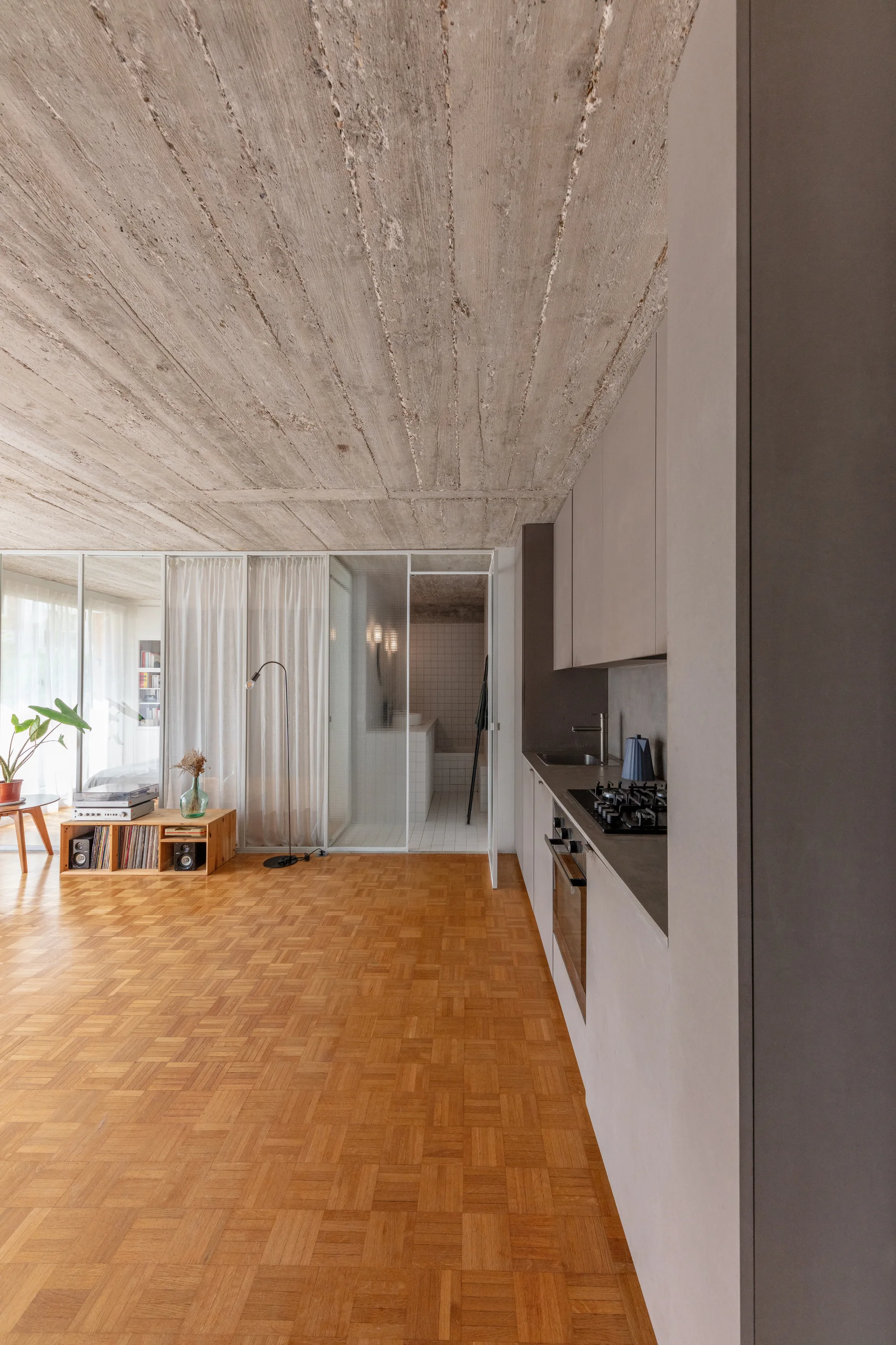 A modern open interior features sleek matte grey kitchen cabinetry, classic wood parquet flooring, and a rough-textured exposed concrete ceiling.