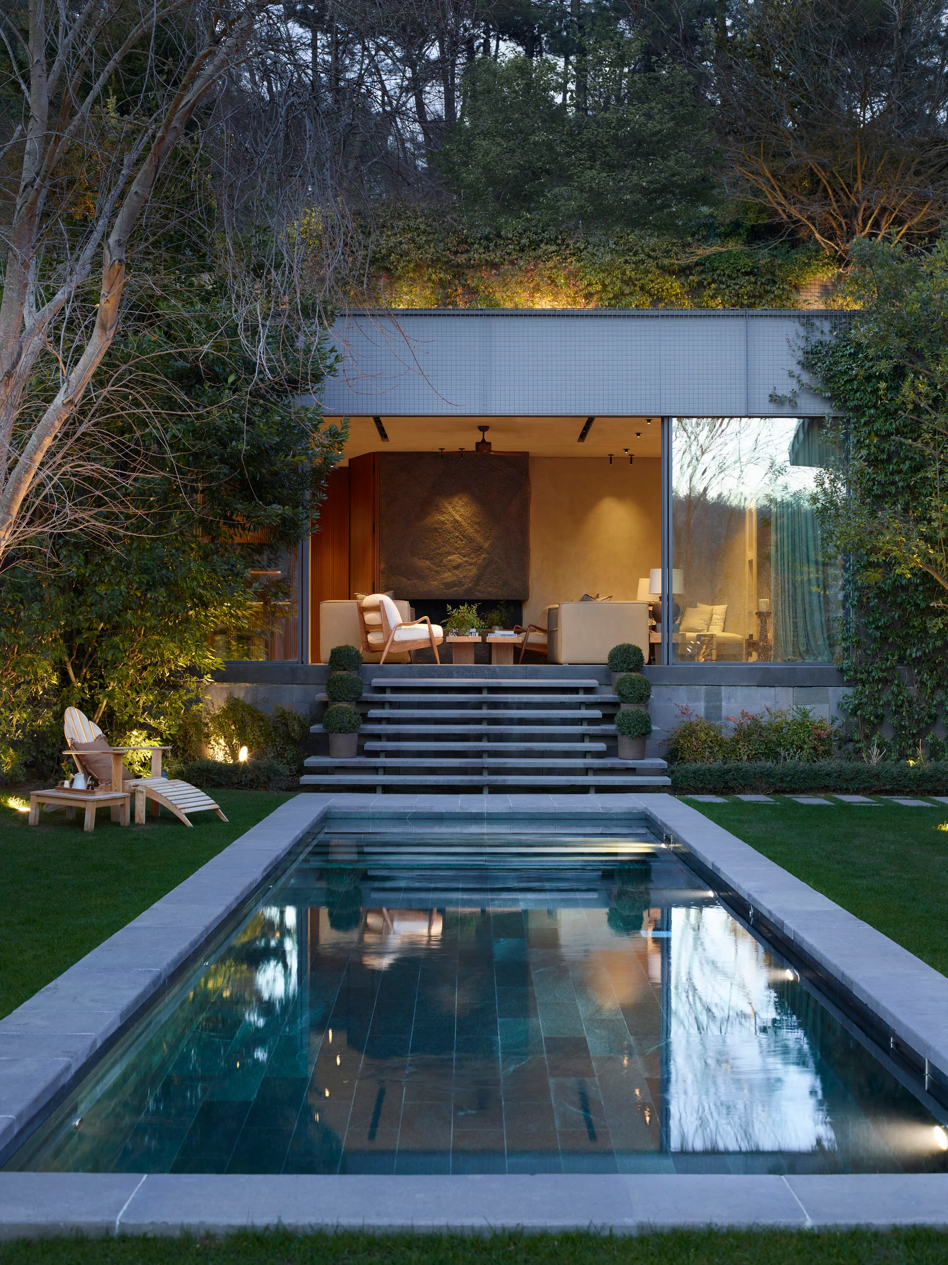 A rectangular swimming pool leads to a modern poolhouse with wide stone steps, large glass walls, and a glowing interior seating area at dusk.