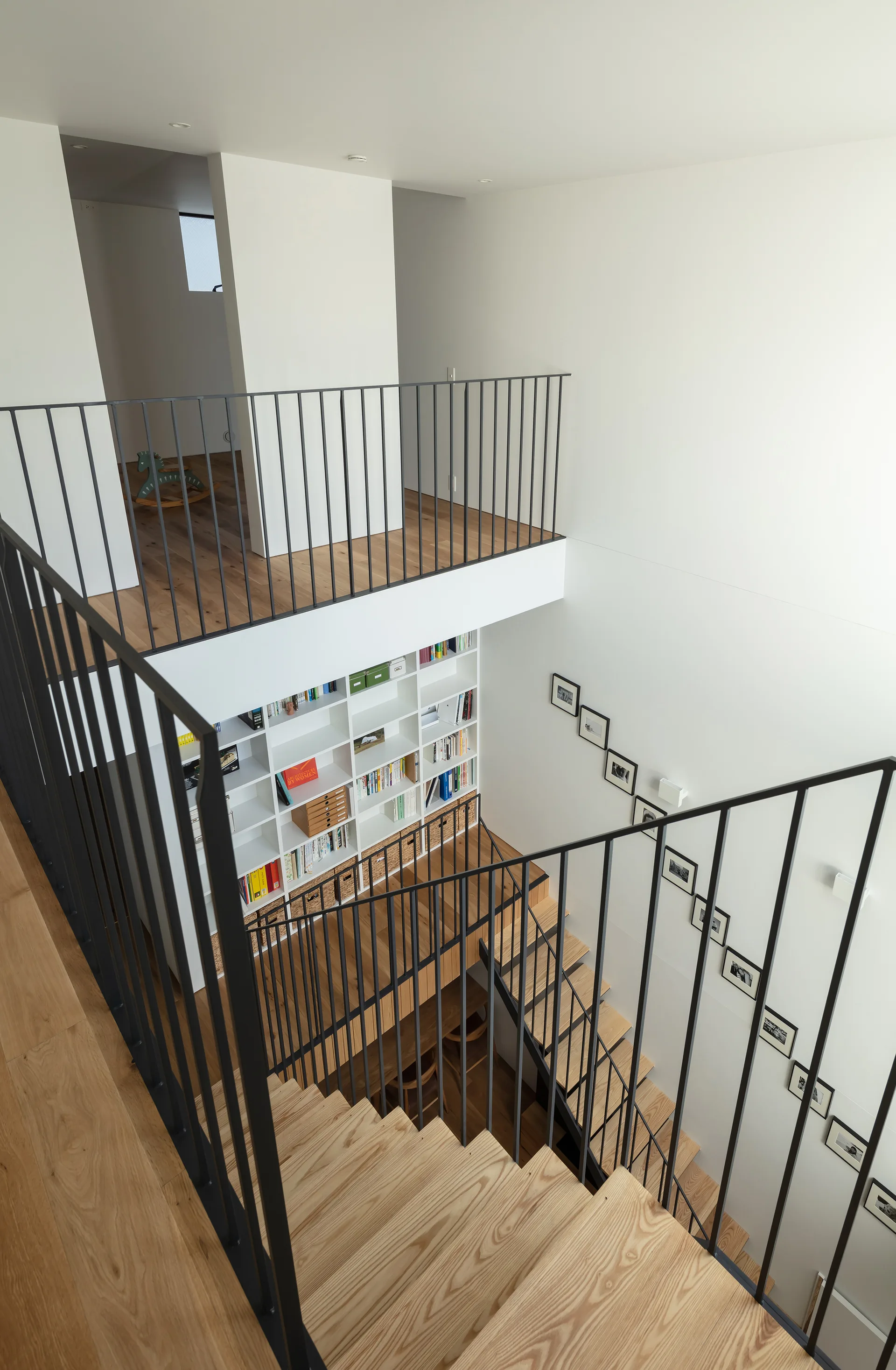 A multi-level wooden staircase with black metal railings features white walls, a large built-in white bookshelf, and a diagonal gallery wall of framed photos.