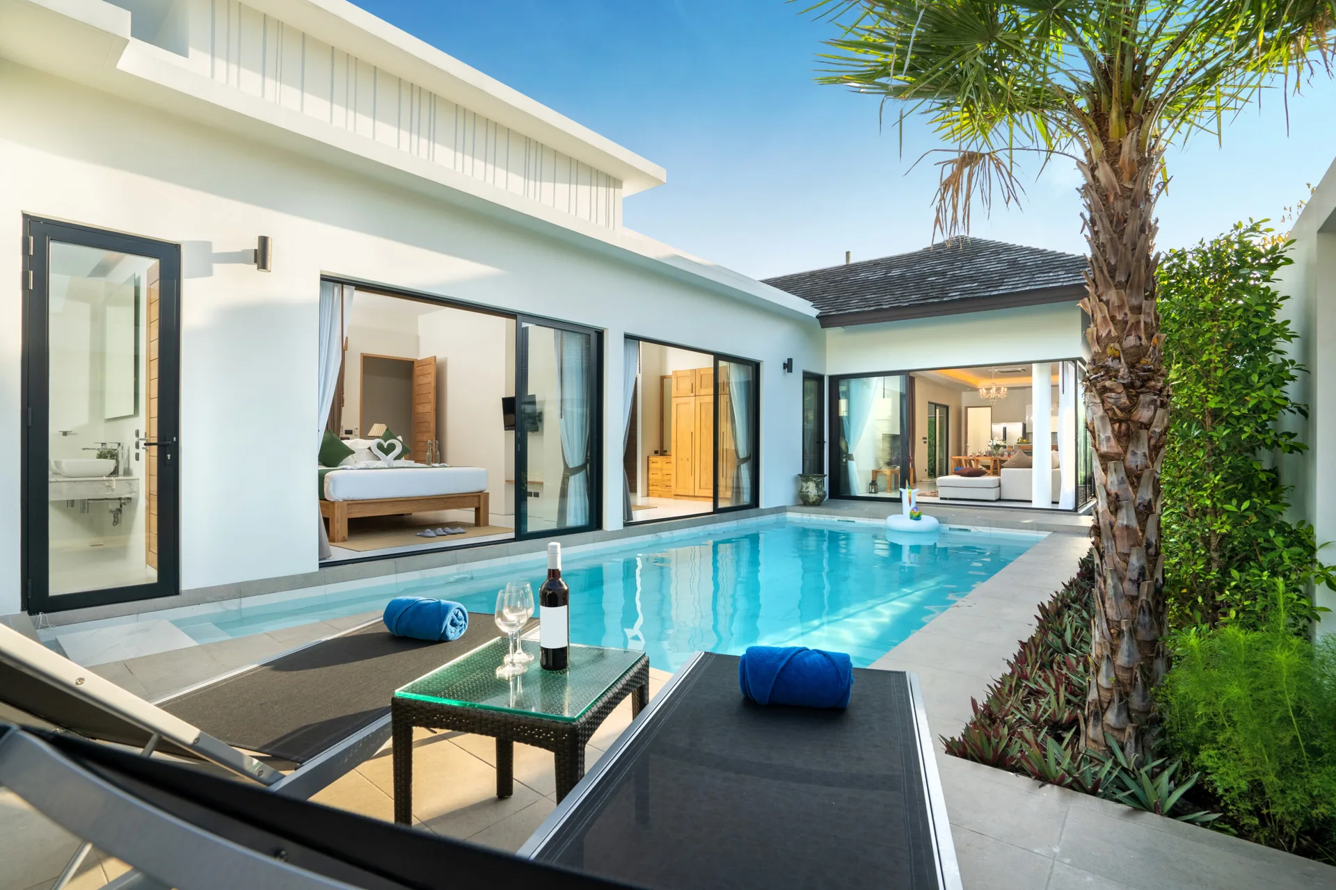 Two black sun loungers sit on a tiled patio next to a turquoise swimming pool, with a modern white villa featuring sliding glass doors in the background.