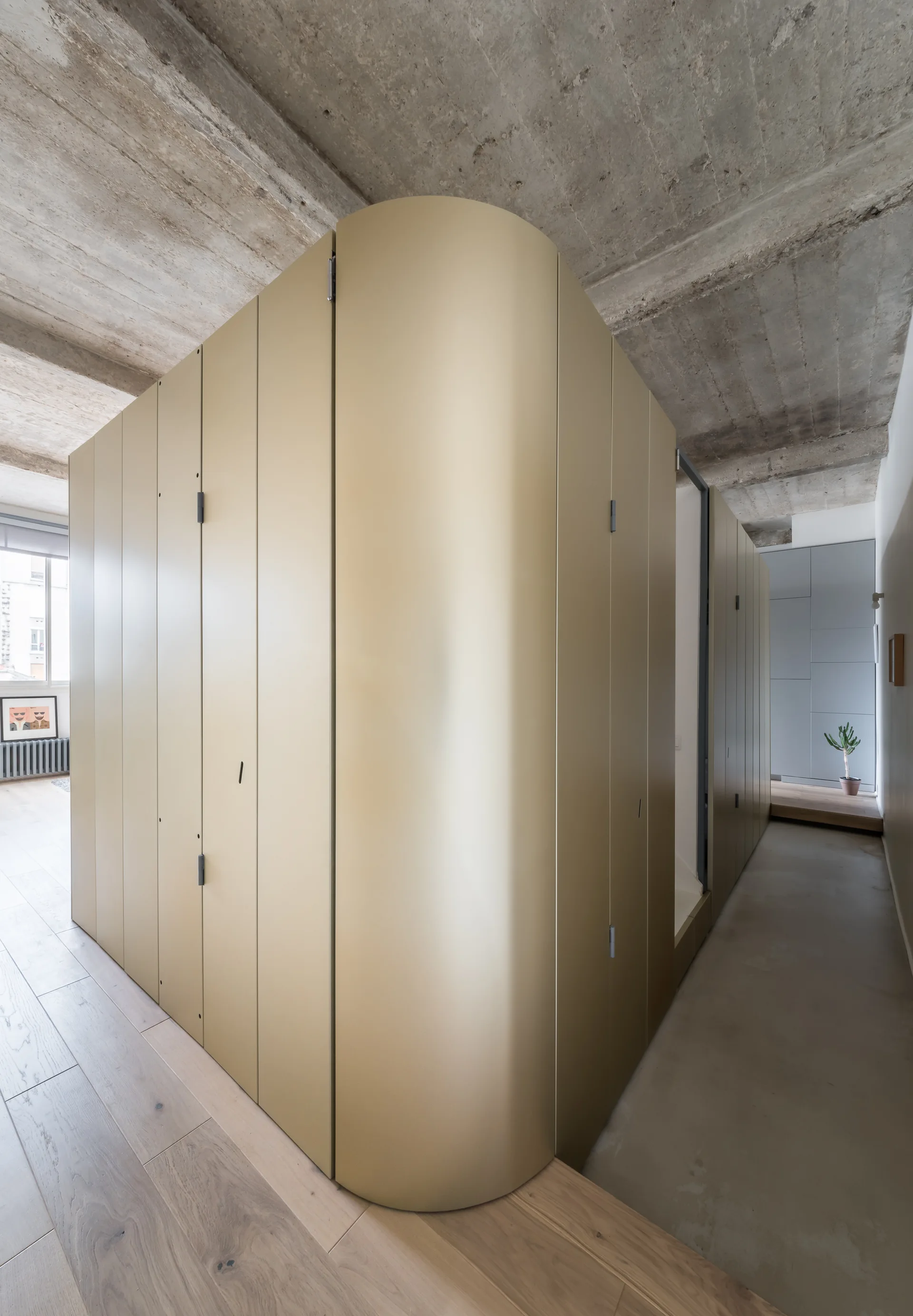 A large curved gold partition cabinet stands under an exposed concrete ceiling in a minimalist space with light wood floors.
