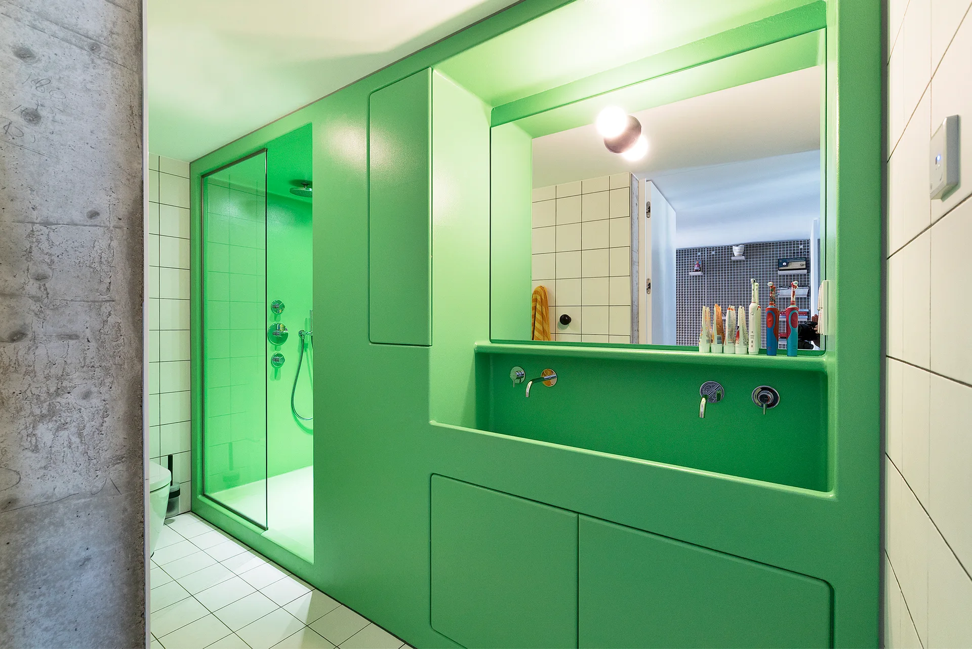 A modern bathroom features a seamless bright green integrated vanity and shower unit, white square tiles, and an exposed concrete wall.