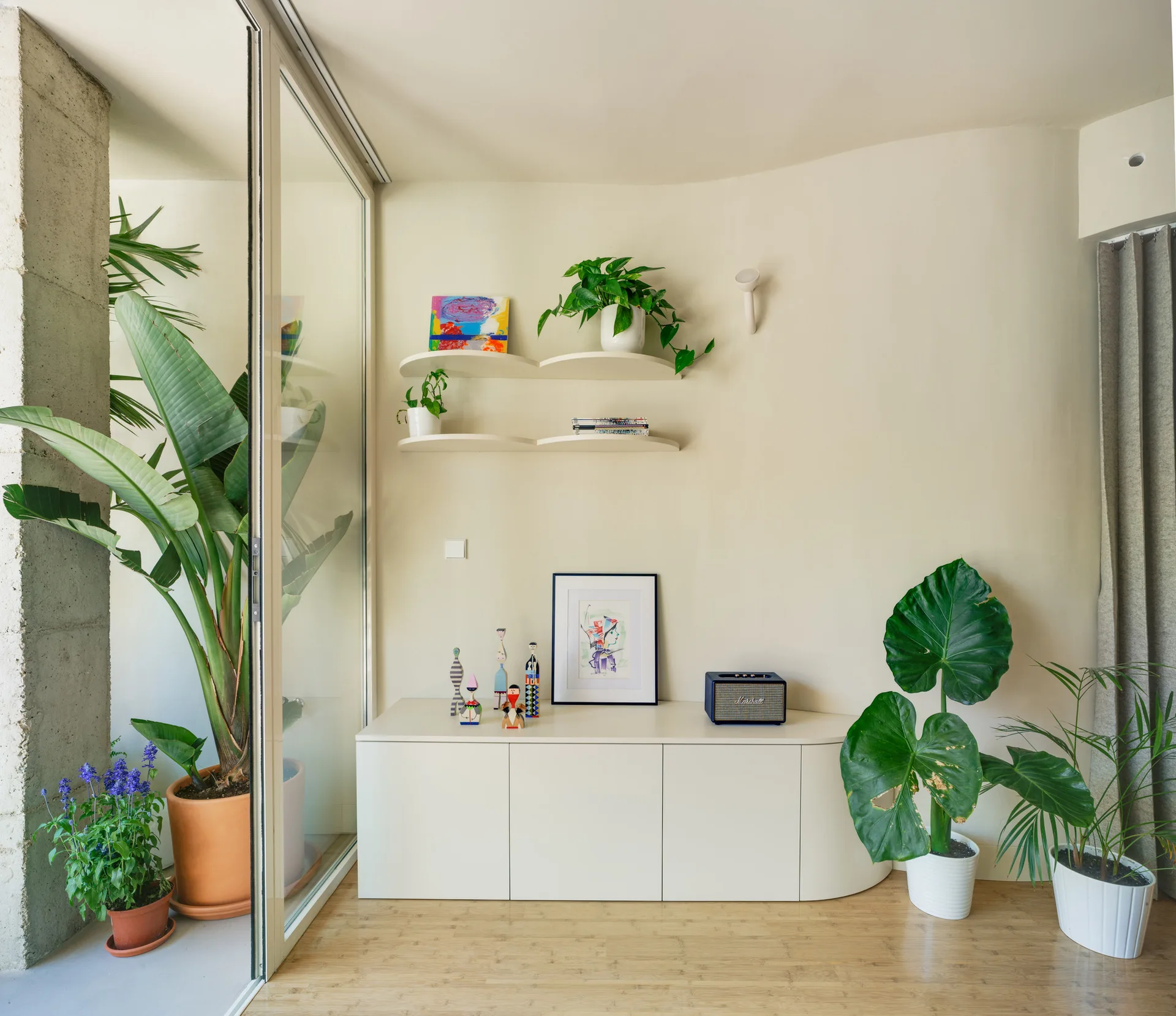 A minimalist cream-colored living room features a low white storage cabinet, curved floating shelves with plants, and a sliding glass door.