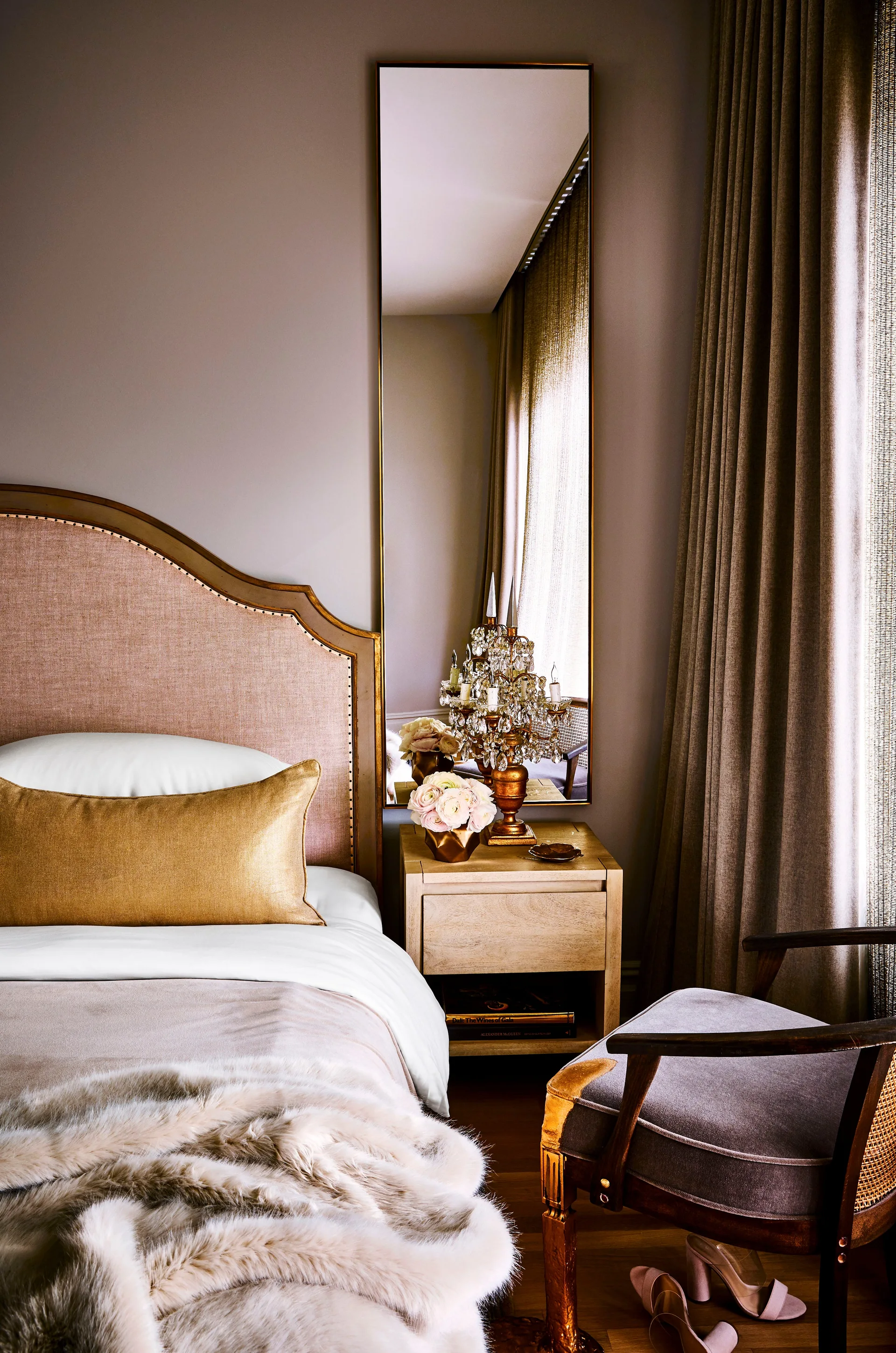 A bedroom corner featuring an upholstered beige headboard, a wooden nightstand with a crystal table lamp, and a tall rectangular wall-mounted mirror.
