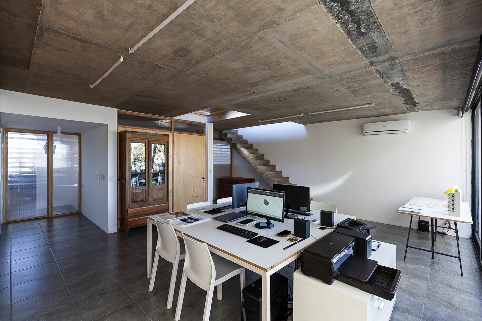 An industrial home office featuring a central white workstation desk, raw concrete ceilings, grey tiled flooring, and a concrete staircase.