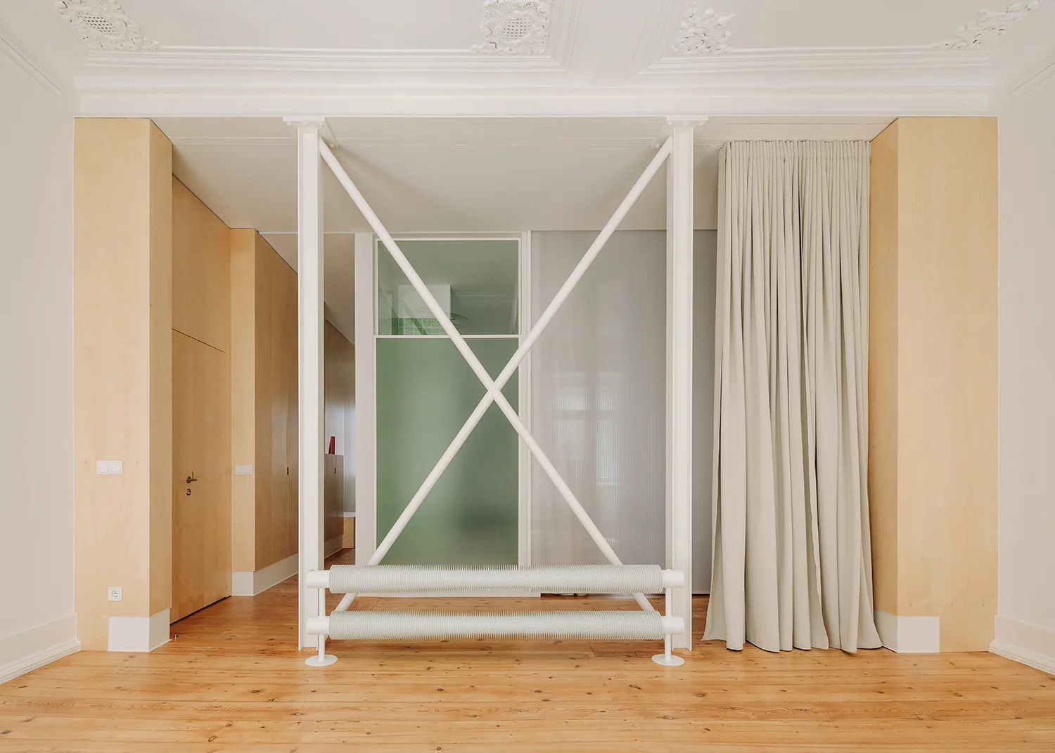 An interior space features wood plank floors, light wood wall panels, a white metal structural x-brace, and an industrial finned radiator.