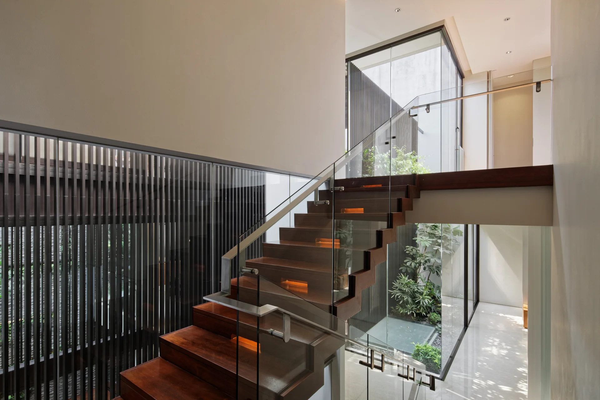 A modern wooden staircase with glass railings and built-in step lights rises past a glass-enclosed indoor garden with green plants.