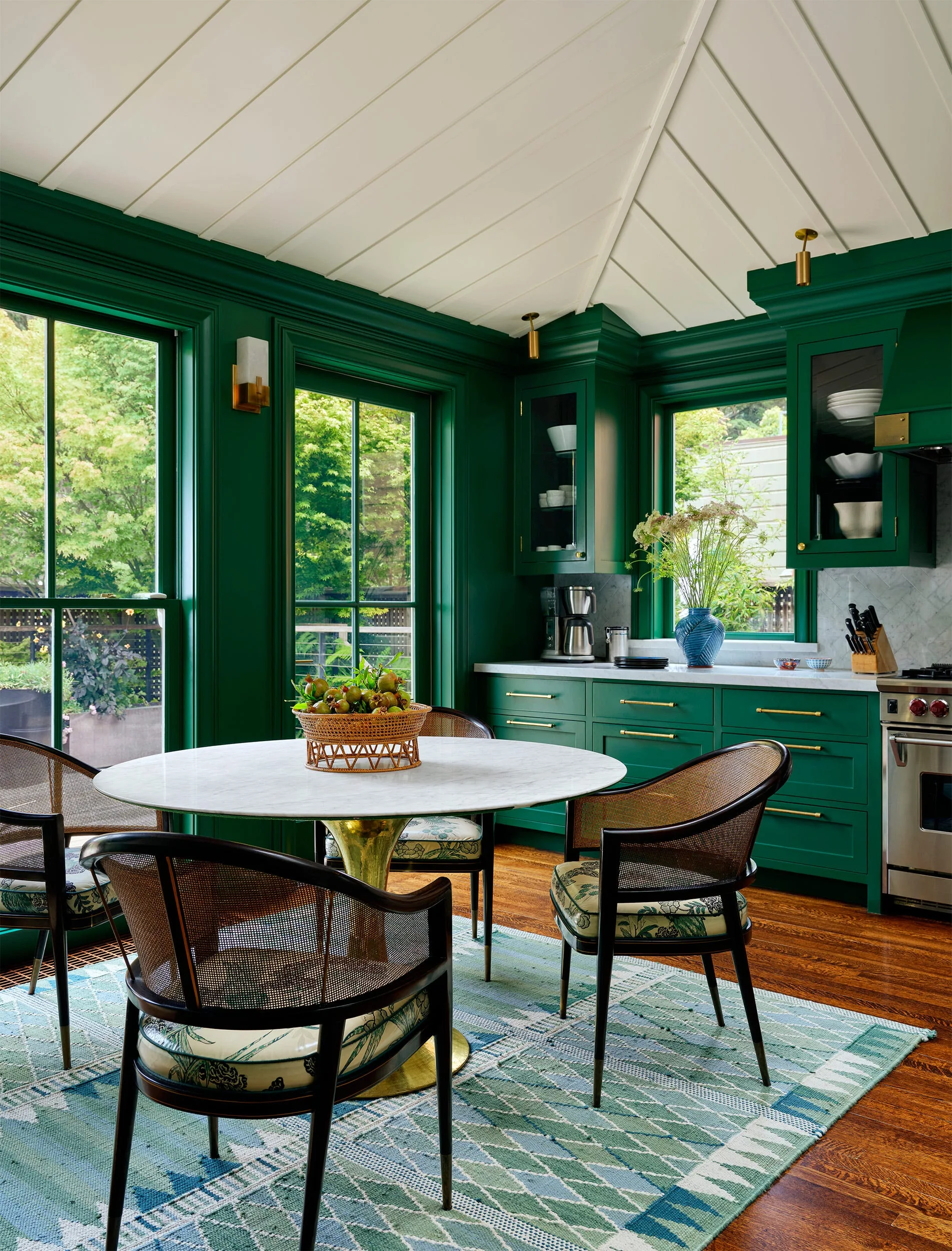 An emerald green kitchen dining nook features a round marble table, cane-back chairs, and a patterned rug beneath a white paneled ceiling.