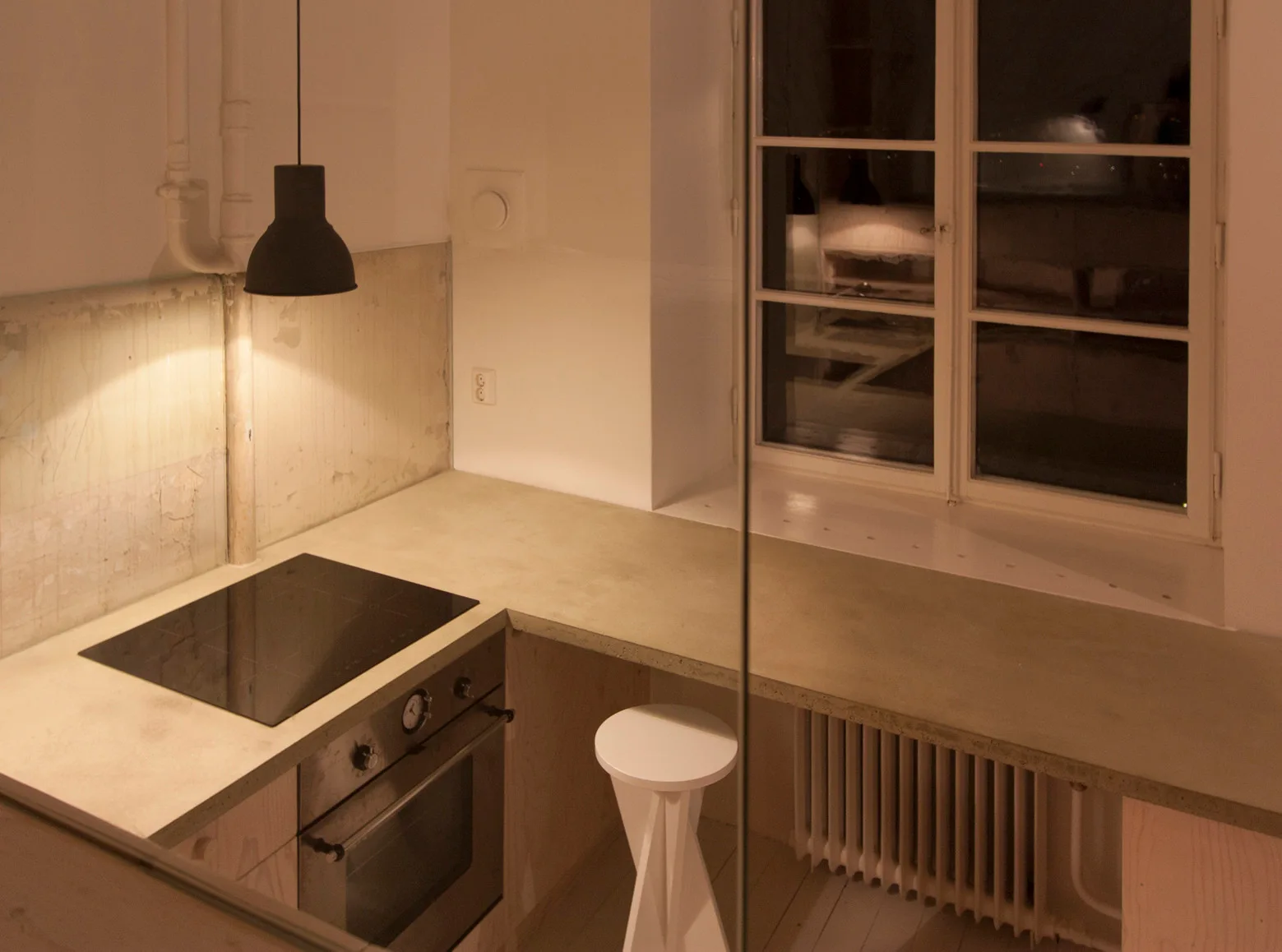 A minimalist kitchen featuring a smooth concrete countertop, integrated oven, white geometric stool, and a black pendant light hanging above the stove.