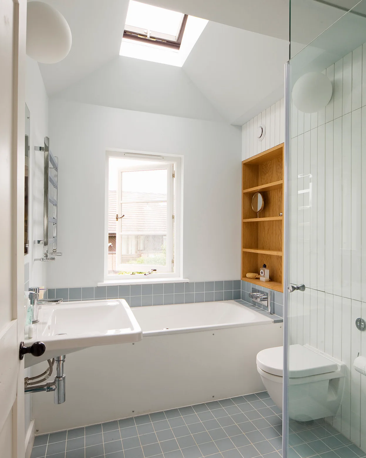 A bright bathroom with light blue floor tiles, a white bathtub, a wall-mounted sink, and a recessed wood shelving unit below a skylight.