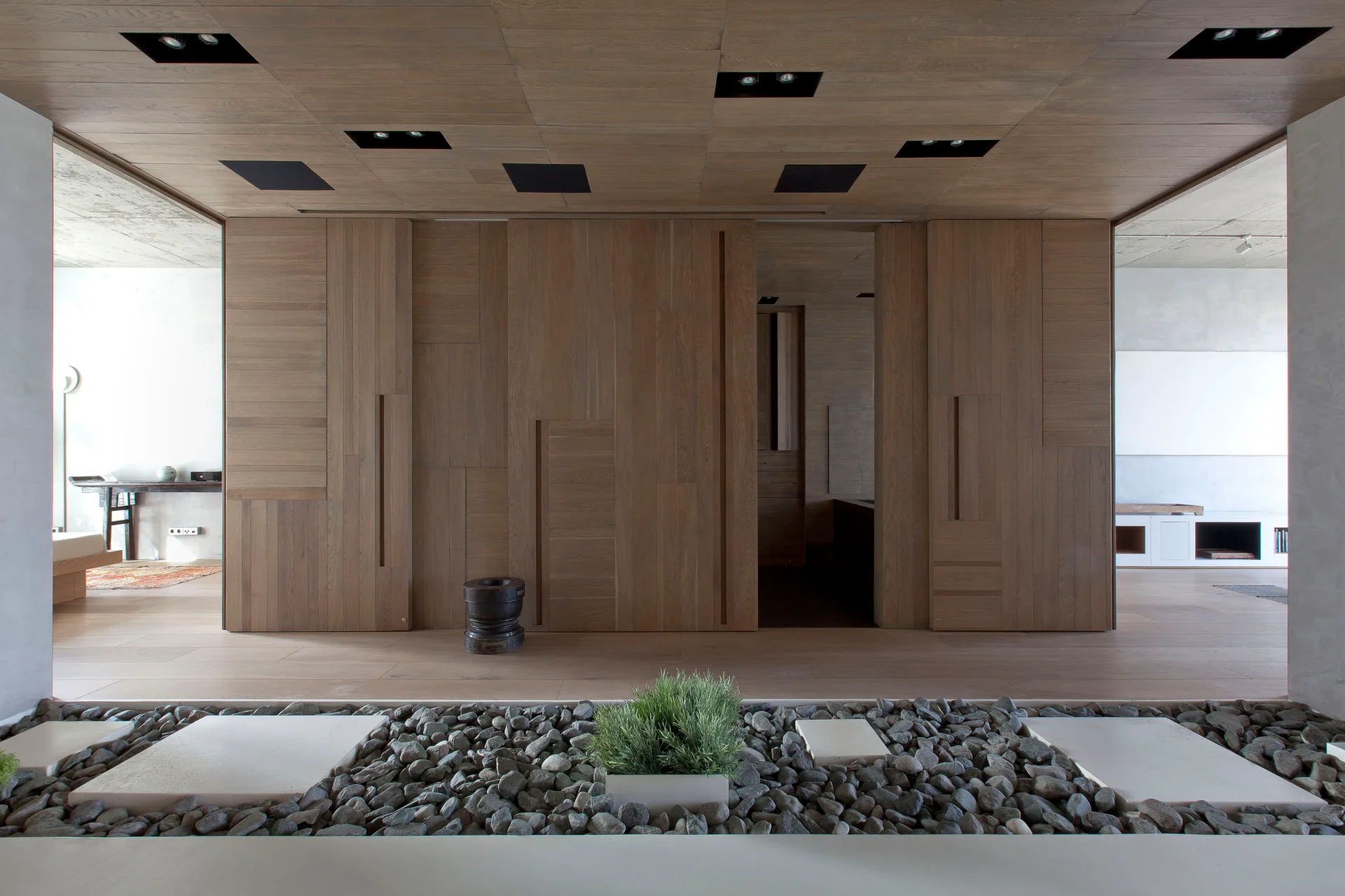 A modern interior space features matching wood paneling on the walls and ceiling, integrated sliding doors, and a bed of river rocks in the foreground.