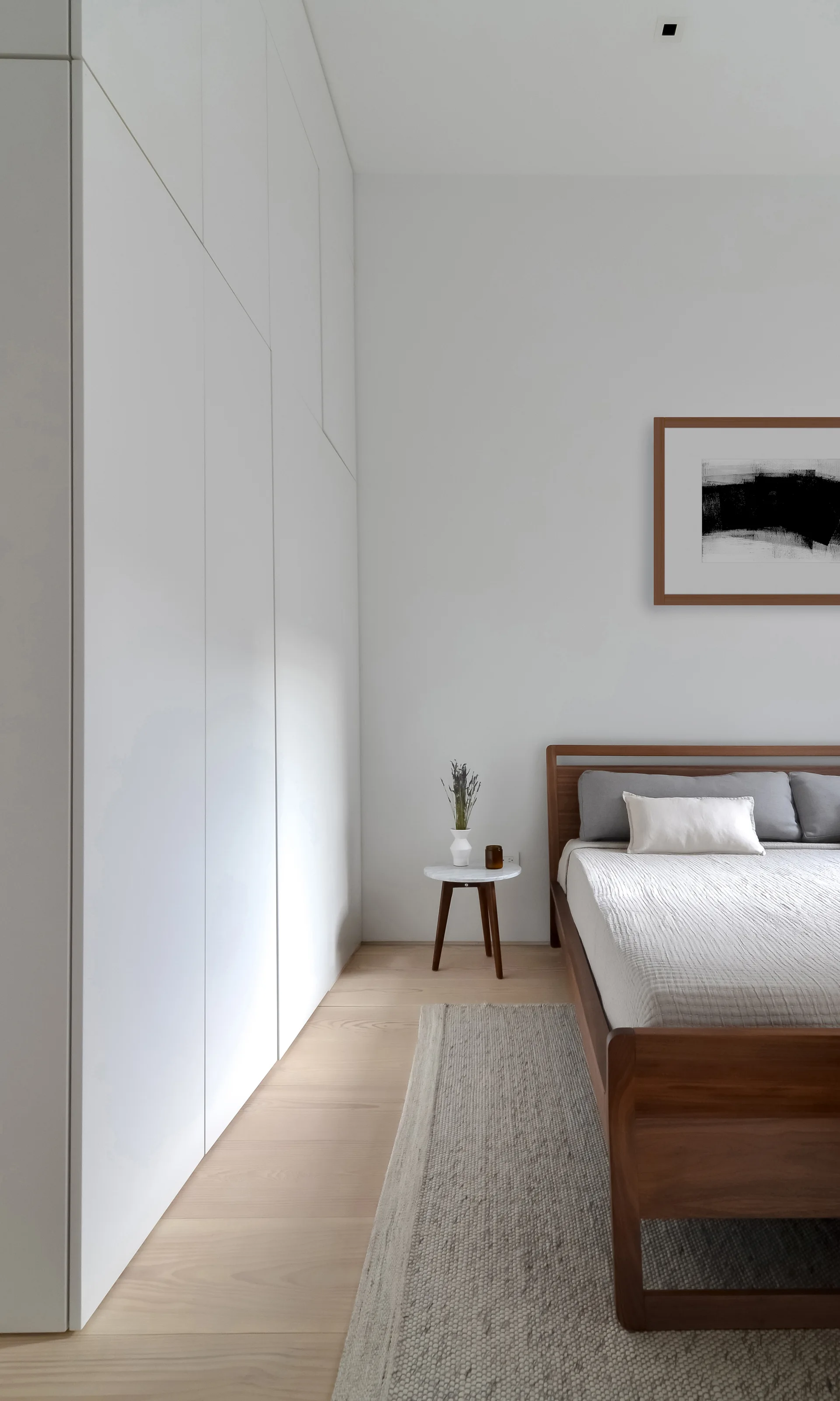 A minimalist bedroom featuring a wooden bed frame, a white built-in wardrobe, a small wood side table, and a textured area rug.