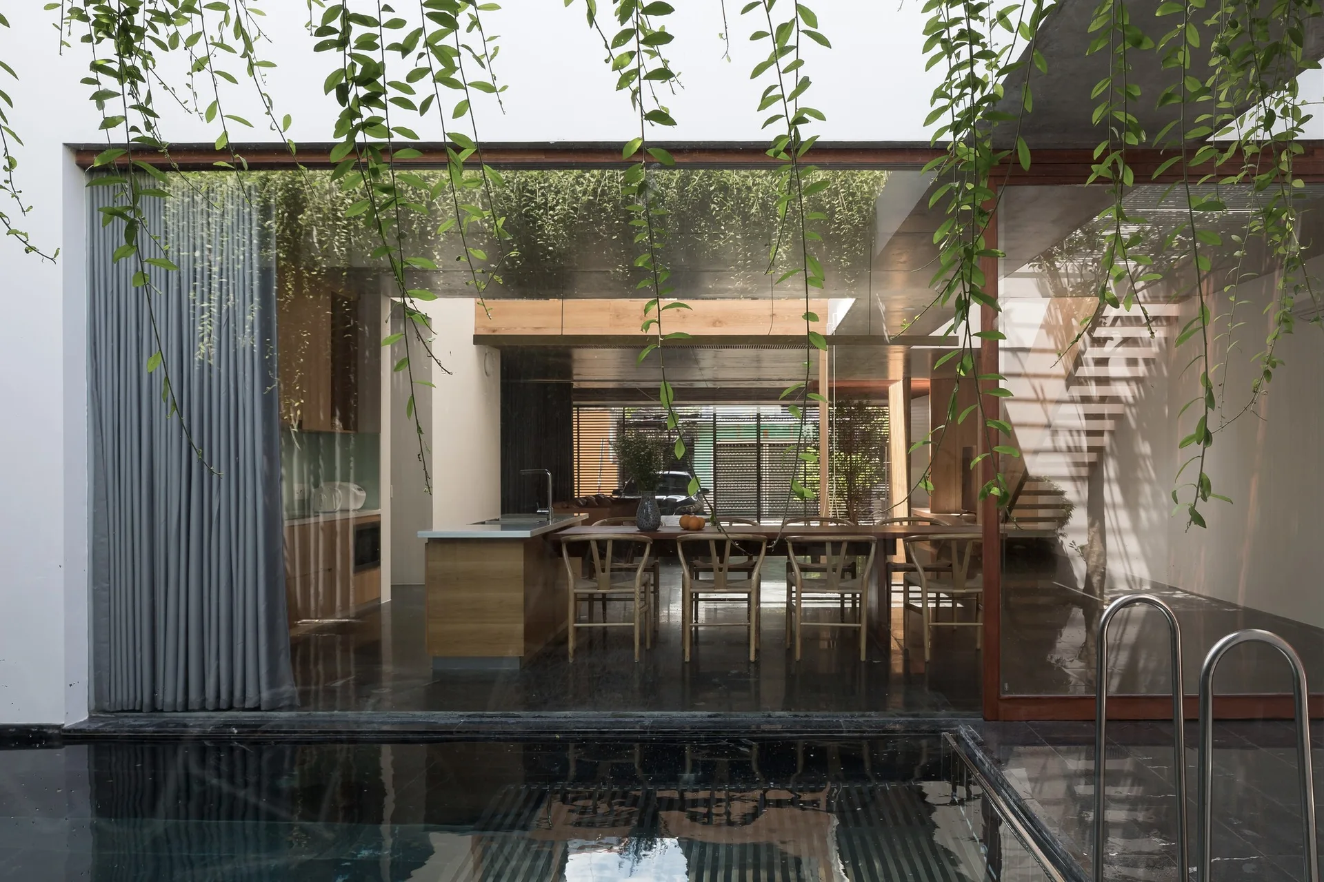 An indoor kitchen and dining area with a wood island and dining chairs is viewed through a glass wall from a dark outdoor pool.