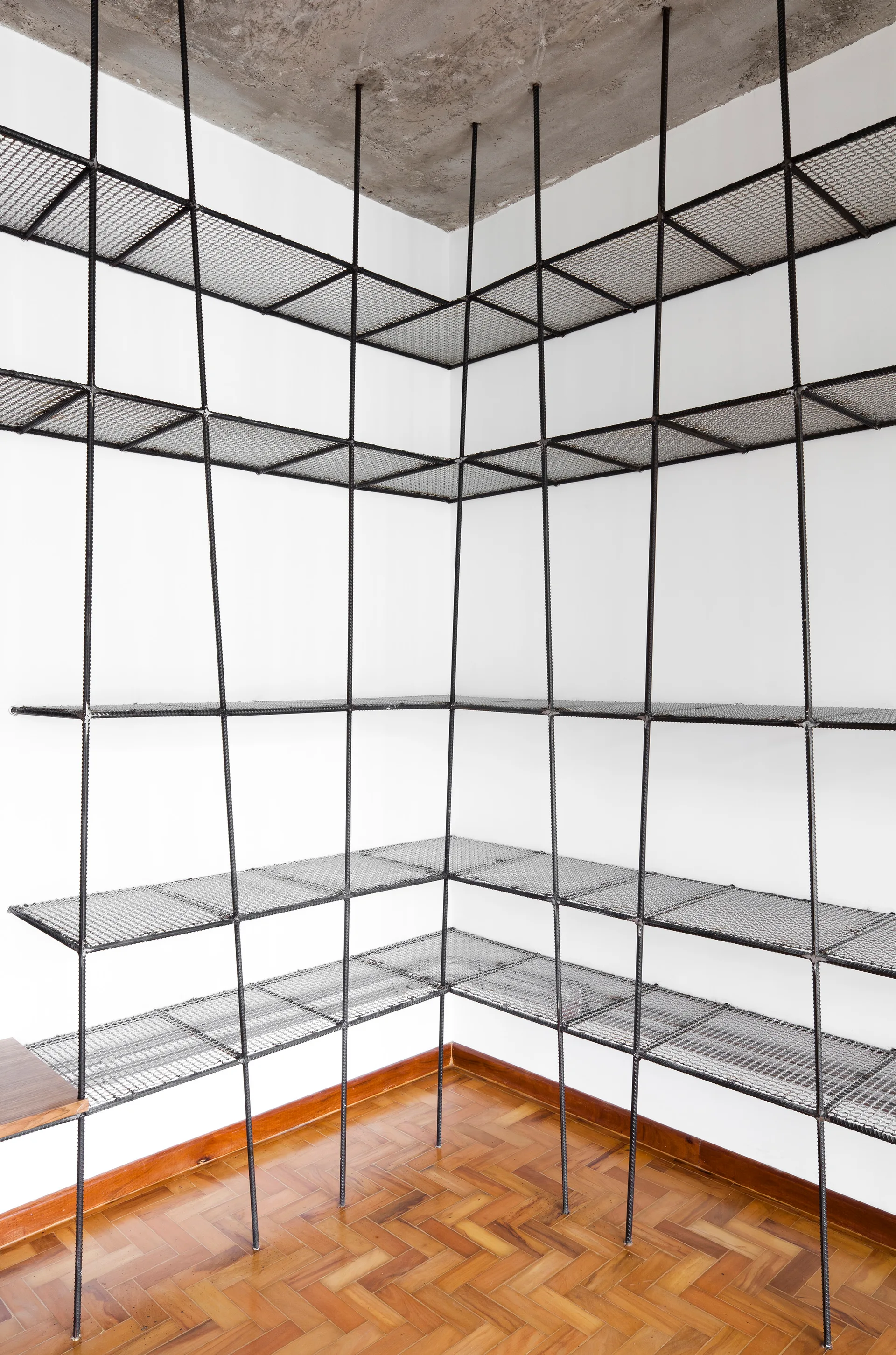 A custom industrial metal wire mesh corner shelving unit stands against white walls under an exposed concrete ceiling with parquet wood flooring.
