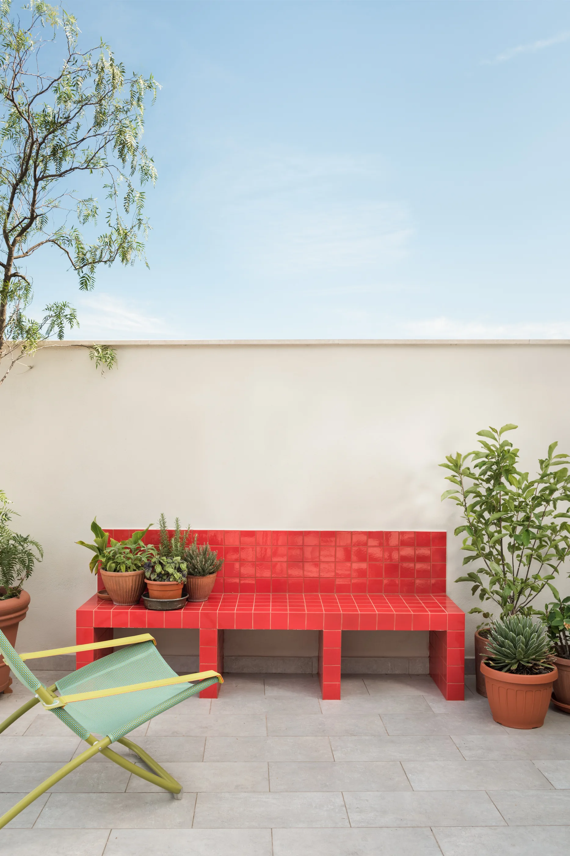 A bright red tiled bench sits against a plain plaster wall on an outdoor patio surrounded by terracotta potted plants and a green folding chair.
