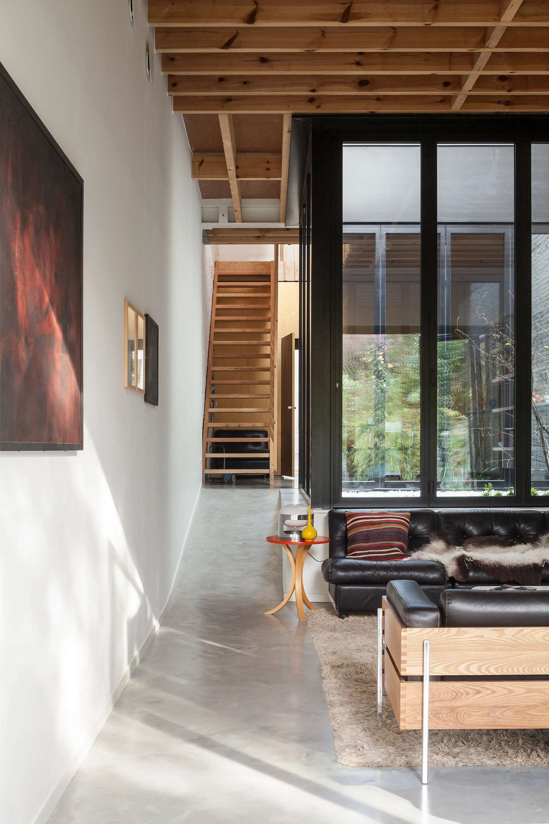 A modern living space features polished concrete floors, exposed wooden ceiling joists, black leather sofas, and a wooden open-riser staircase in the back.