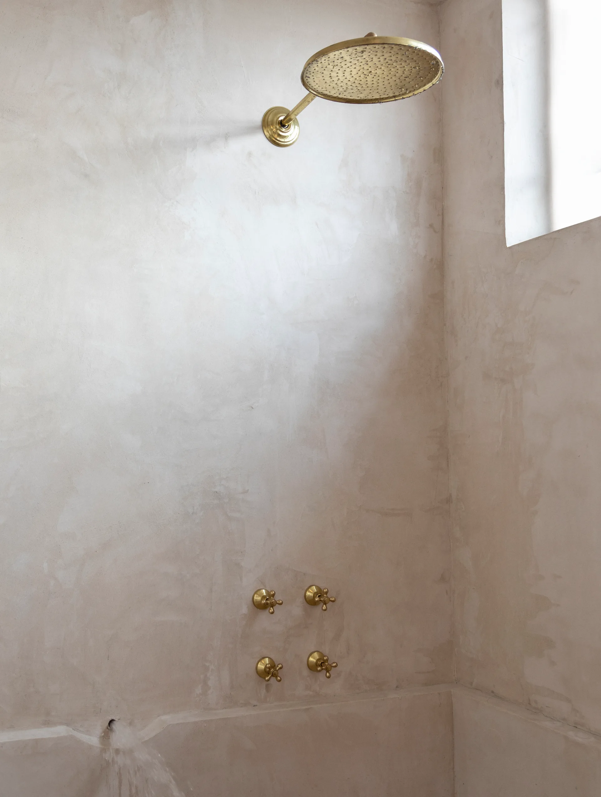 A brass rain shower head and four matching cross-handle knobs are mounted on a beige, textured plaster wall in a shower stall.