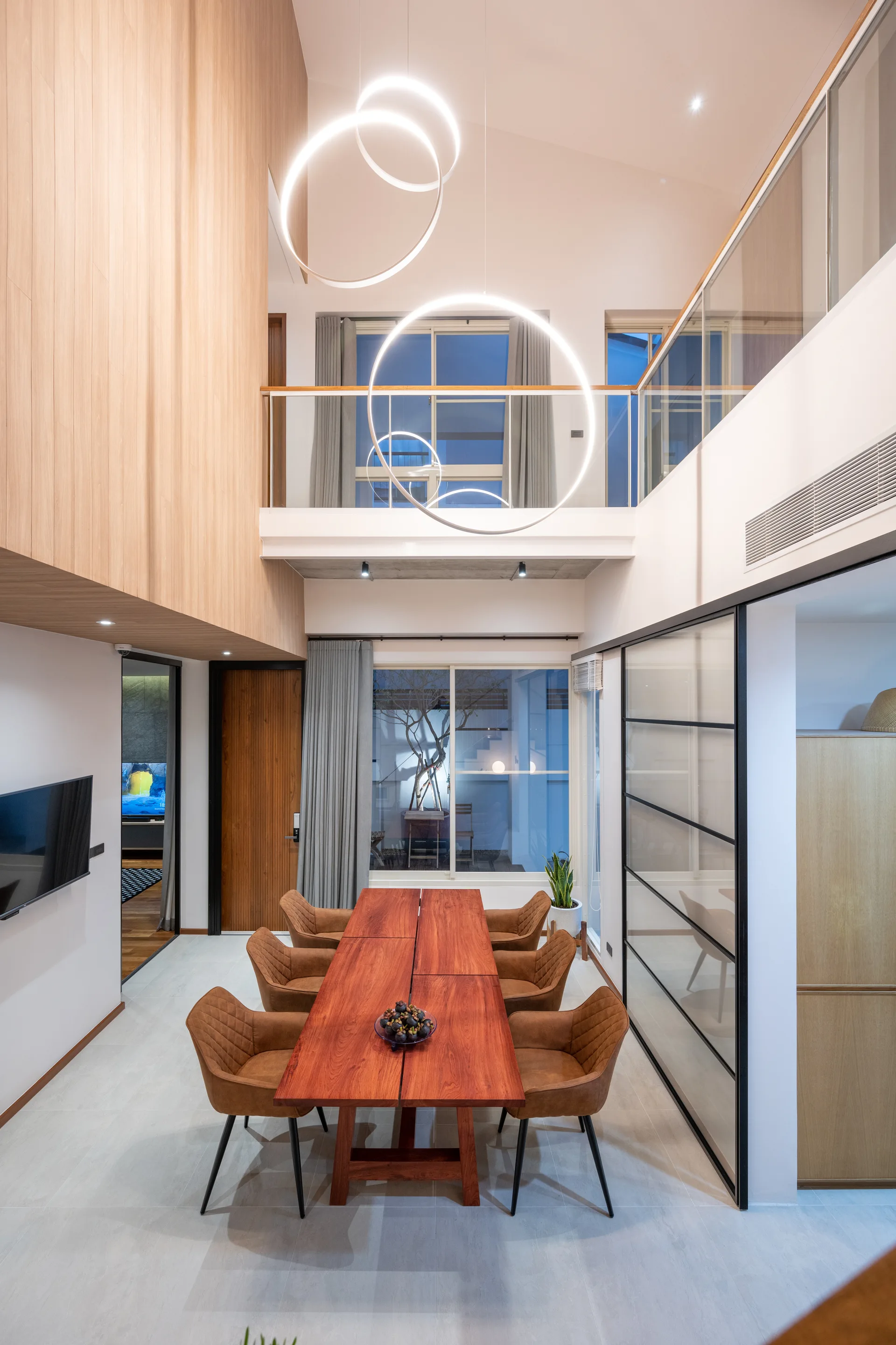 A modern dining room with a long wood table, brown quilted chairs, and large circular LED pendant lights under a double-height ceiling with a mezzanine.