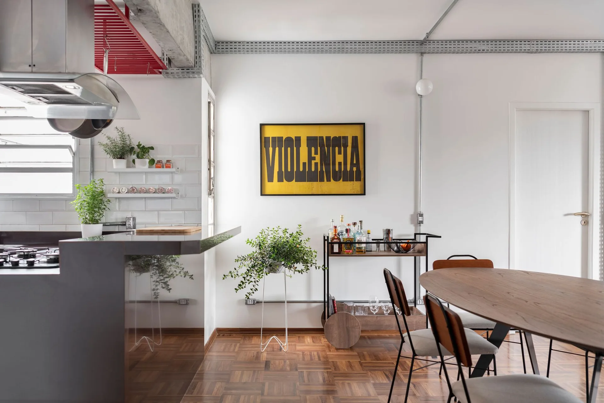 An industrial dining area features an oval wooden table, a mobile bar cart under a yellow framed poster, and exposed metal conduit on white walls.