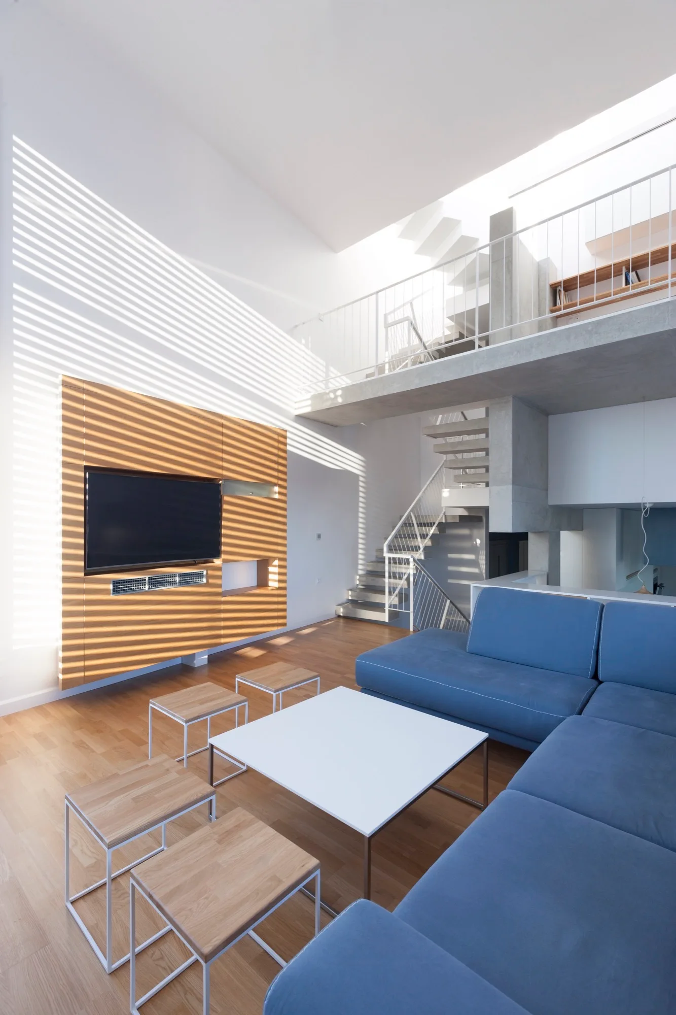 A modern loft living room features a blue sectional sofa, a wood-paneled media wall with a TV, and a white metal staircase leading to a mezzanine.