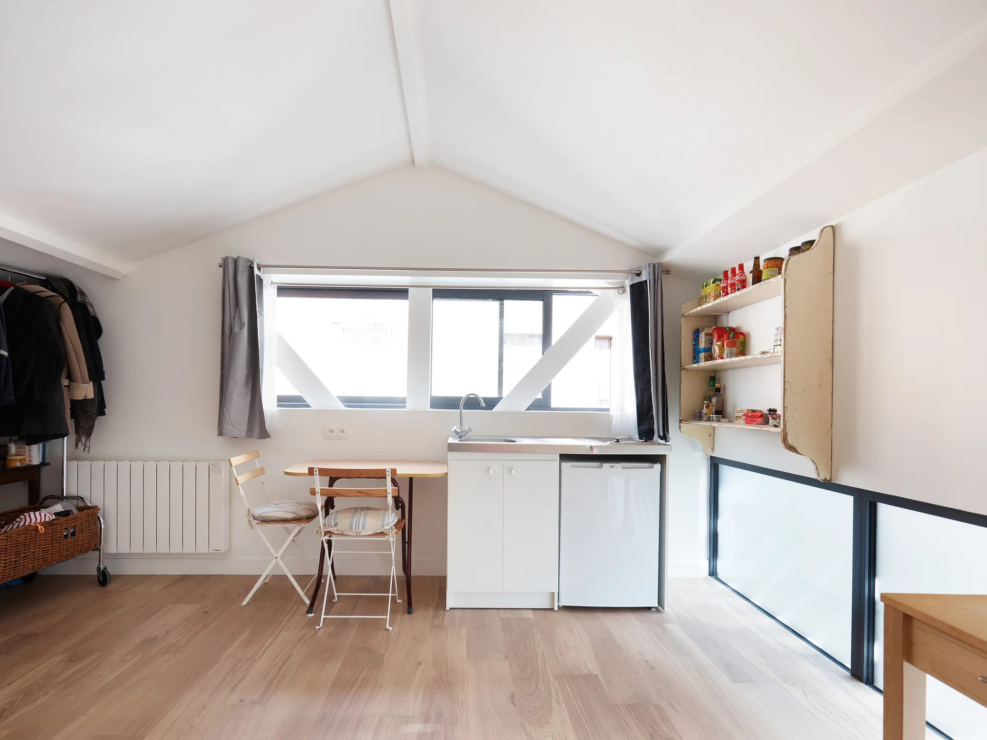 A minimalist kitchenette with a white cabinet and mini-fridge sits under a pitched ceiling beside a small wooden dining table with two folding chairs.