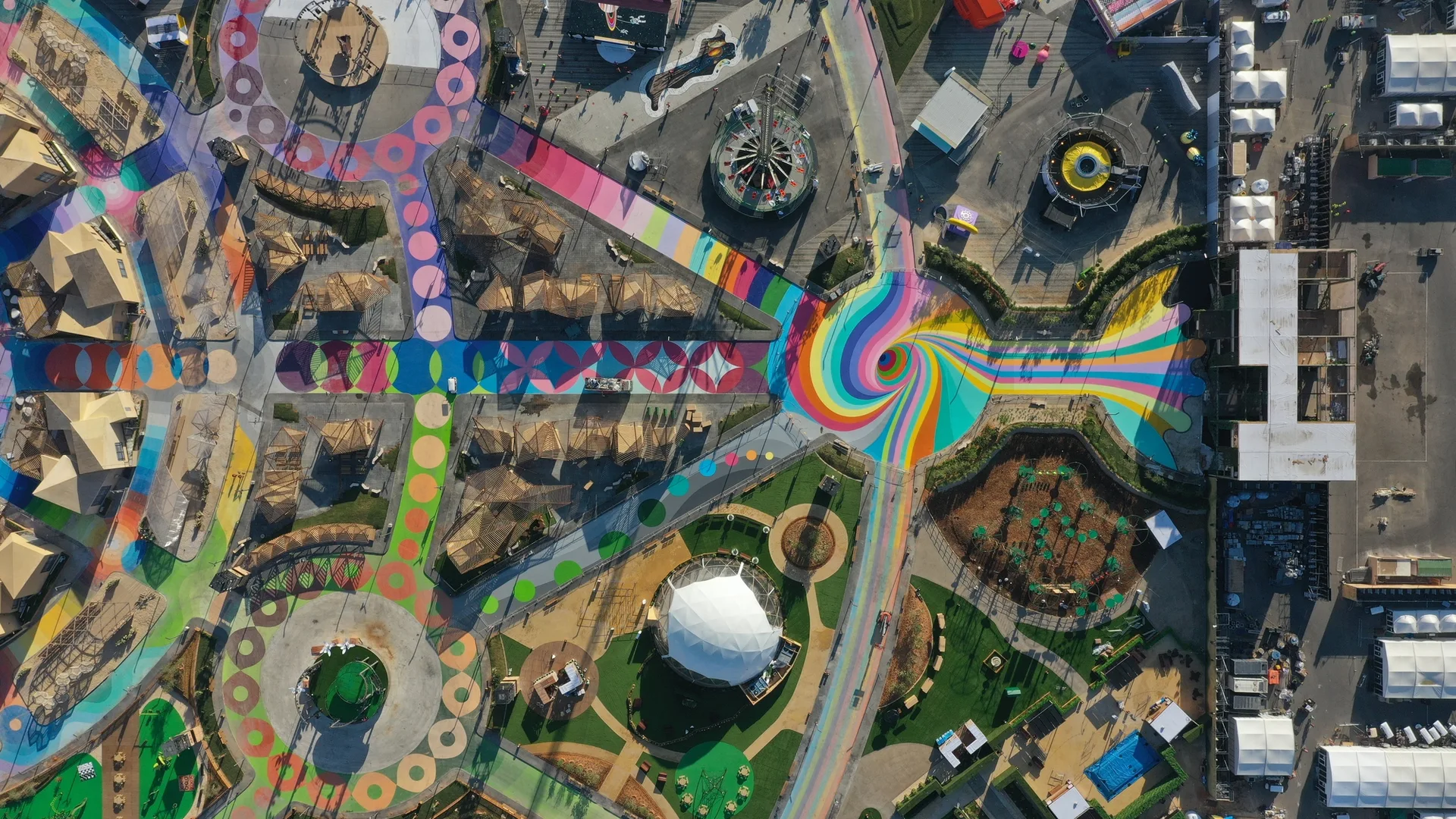 An aerial view of an outdoor park featuring colorful swirling and striped paved pathways, green lawns, and small wooden pavilion structures.