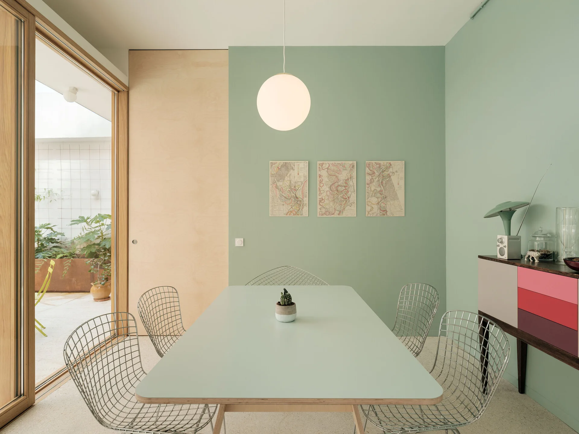 A minimalist dining area features a light green table surrounded by wire chairs, set against a sage green wall next to a large wood-framed sliding glass door.