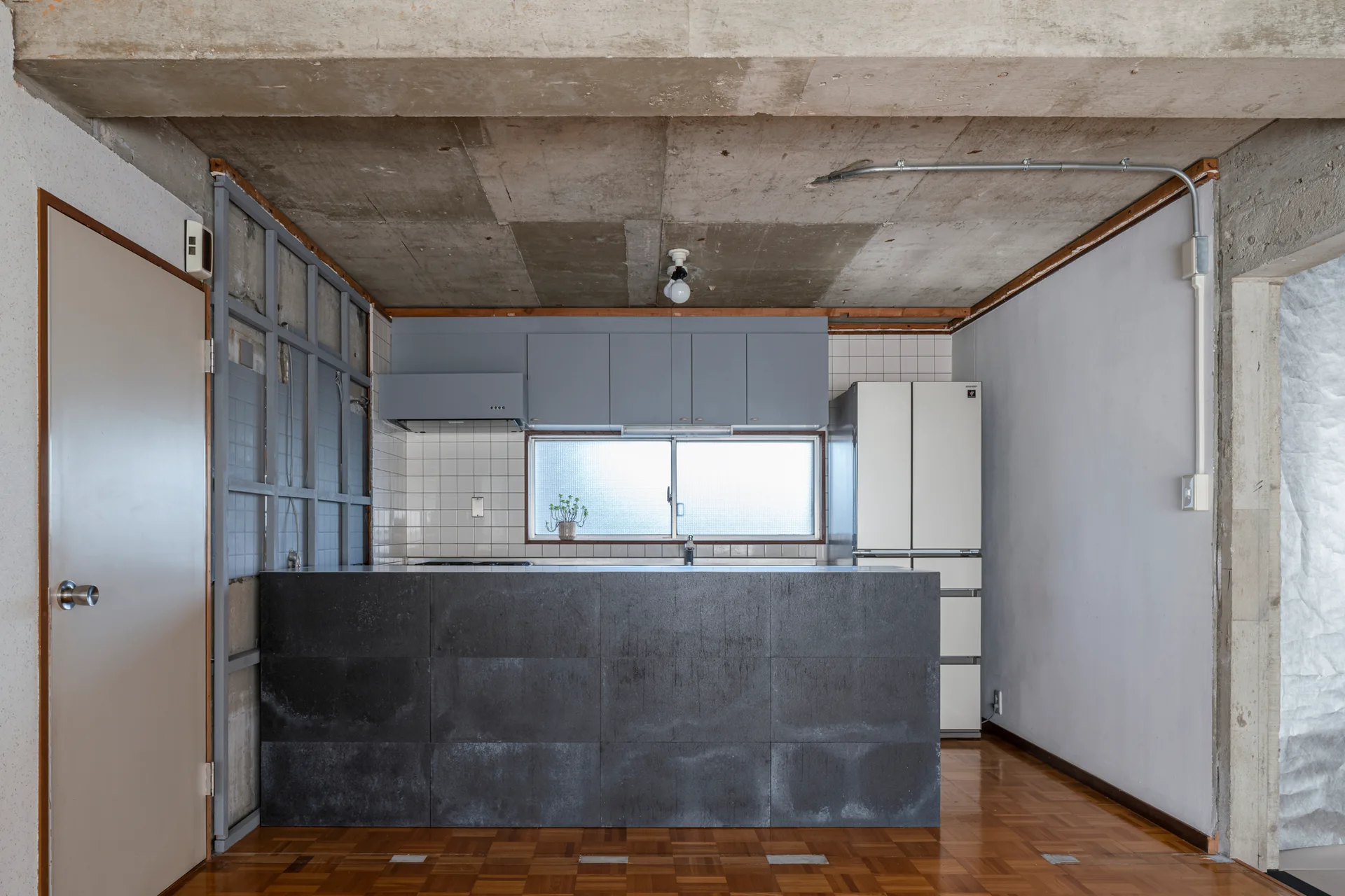 A minimalist kitchen features a dark grey stone-clad island, grey upper cabinetry, an exposed concrete ceiling, and a warm wood parquet floor.
