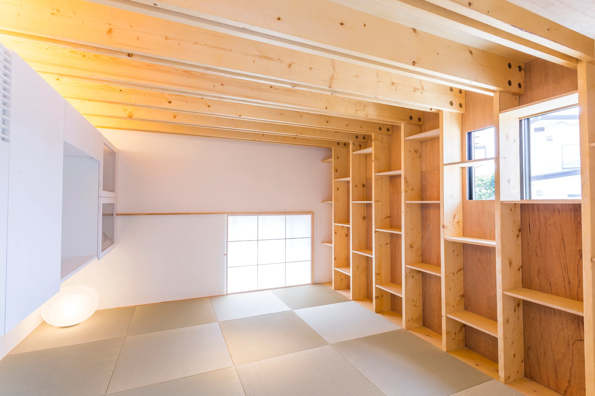 A minimalist room features light wood exposed ceiling beams, built-in wooden wall shelving, a glowing floor lamp, and square tatami mats.